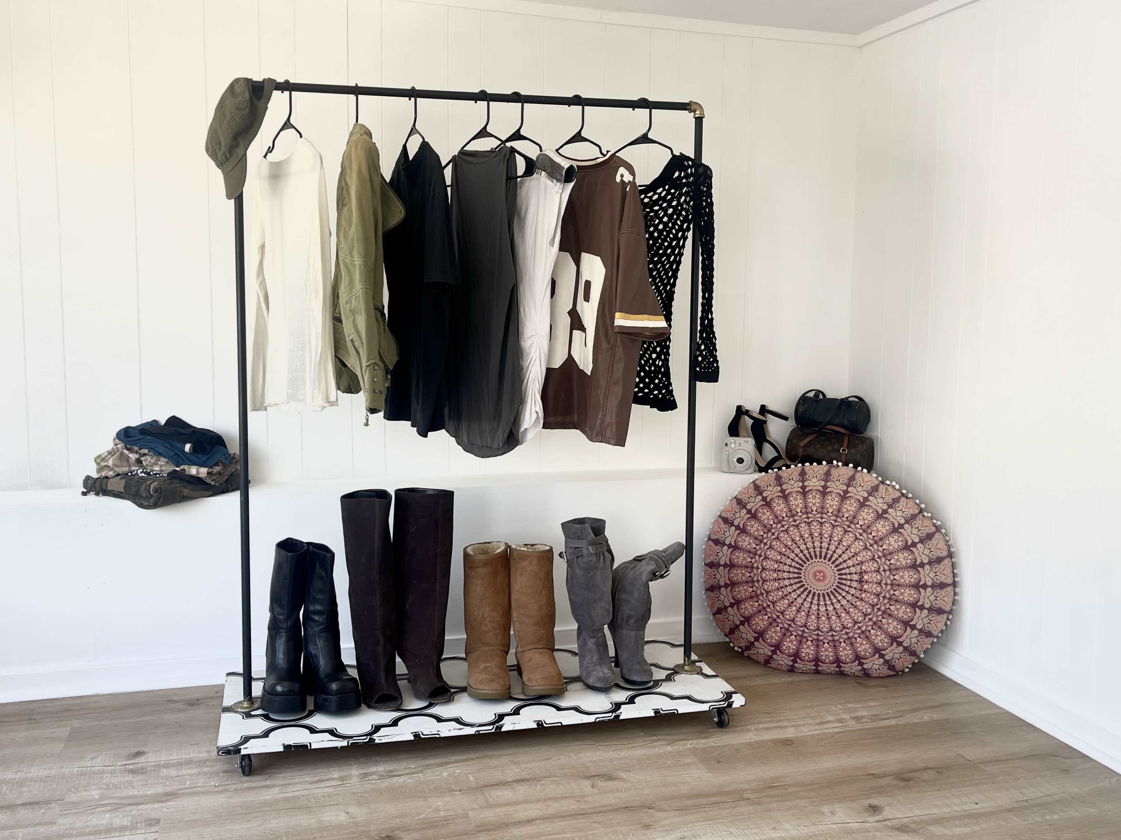 A metal clothing rack displays a variety of garments alongside several pairs of boots and a decorative round rug on a wooden floor.