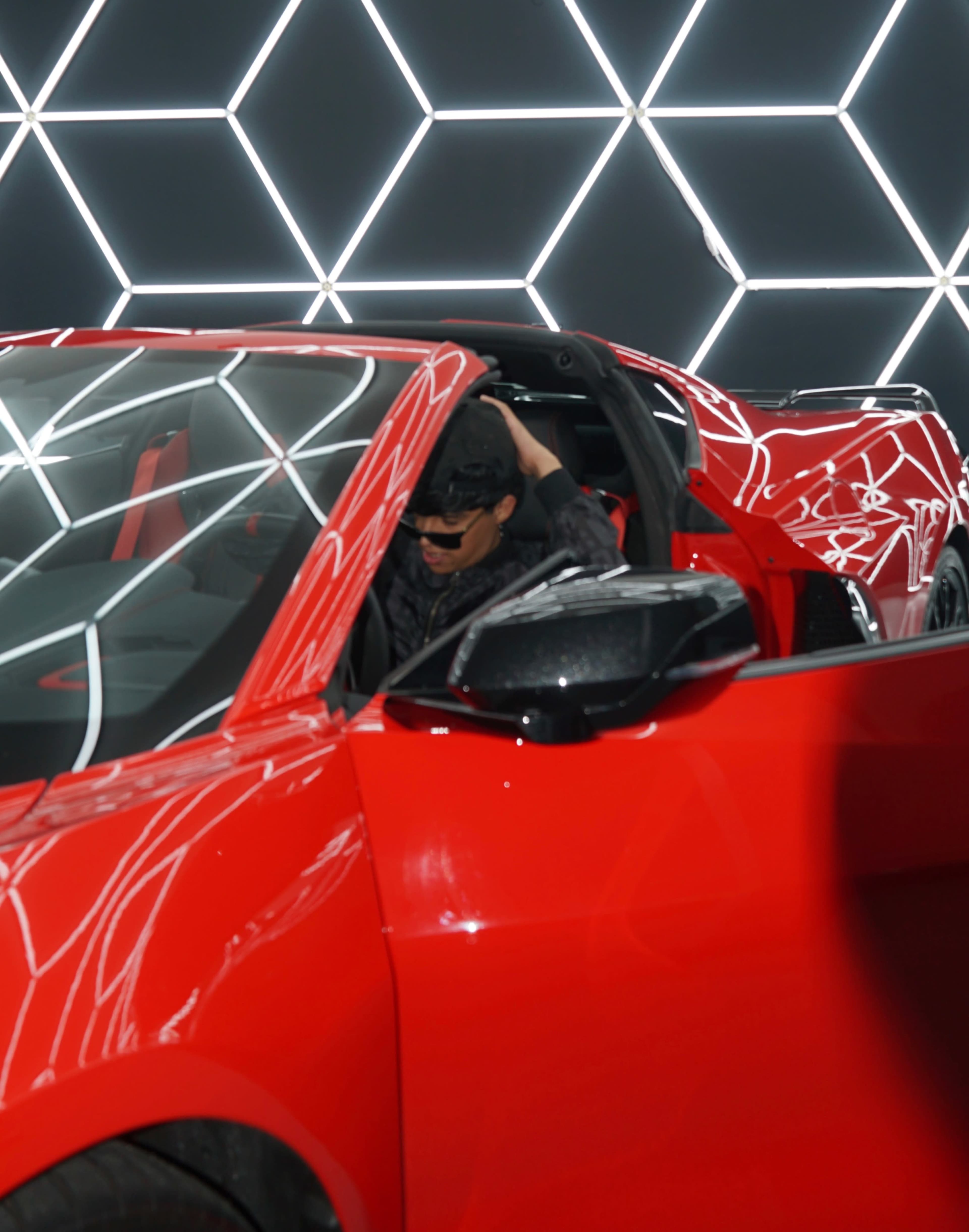 A person in sunglasses is adjusting their hair while seated in a red convertible sports car, with a geometric hexagonal light display in the background.