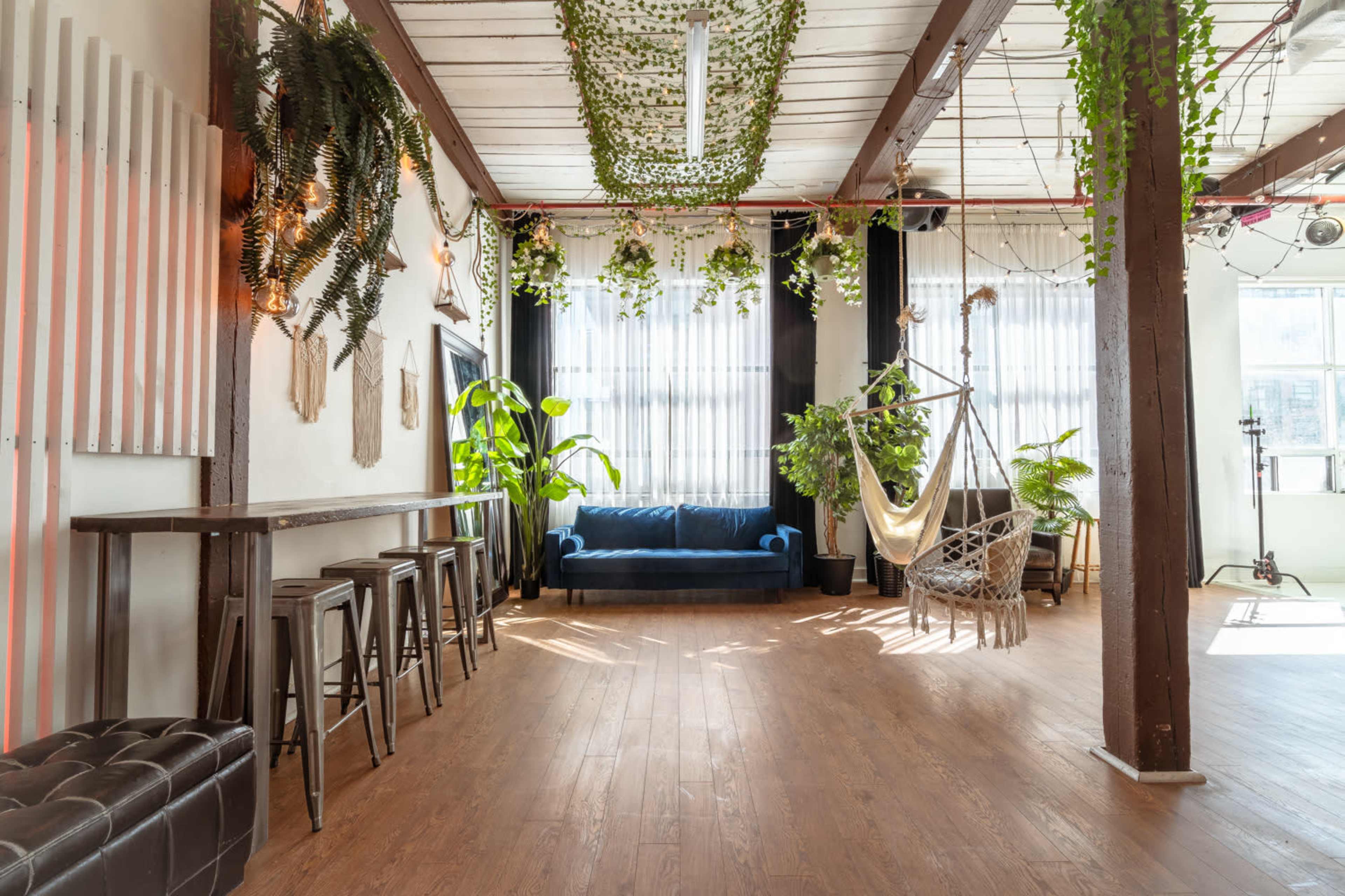 A spacious interior features a blue couch, hanging plants, a swing chair, and a wooden bar with stools.