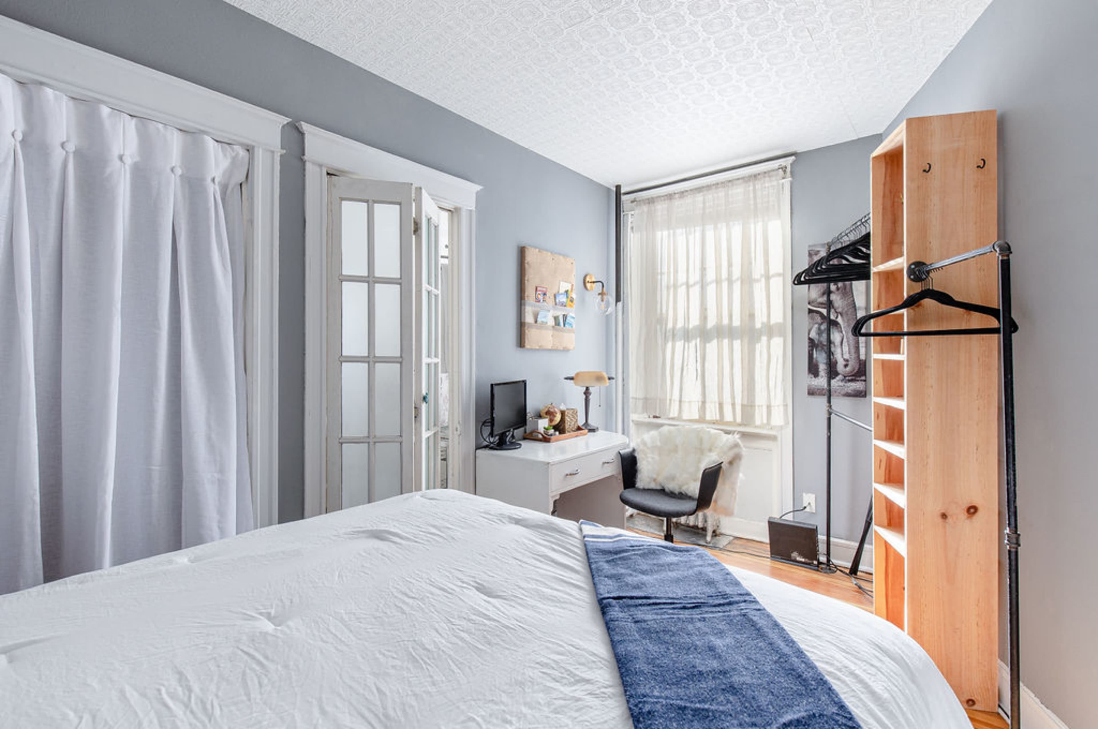 A cozy bedroom featuring a bed with white bedding, a small desk with a chair, a wooden shelving unit, and a large window with sheer curtains.