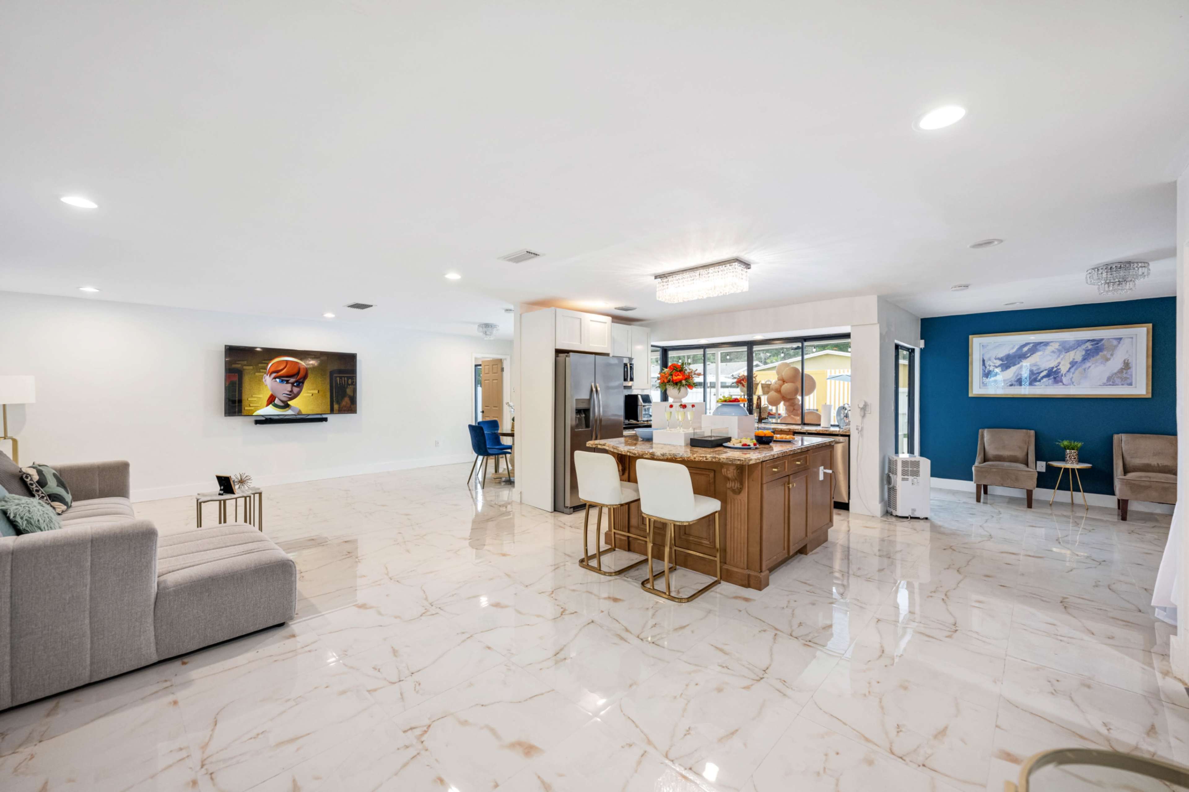 The image shows a spacious, modern living area with a kitchen island, a large television on the wall, and light-colored marble flooring.