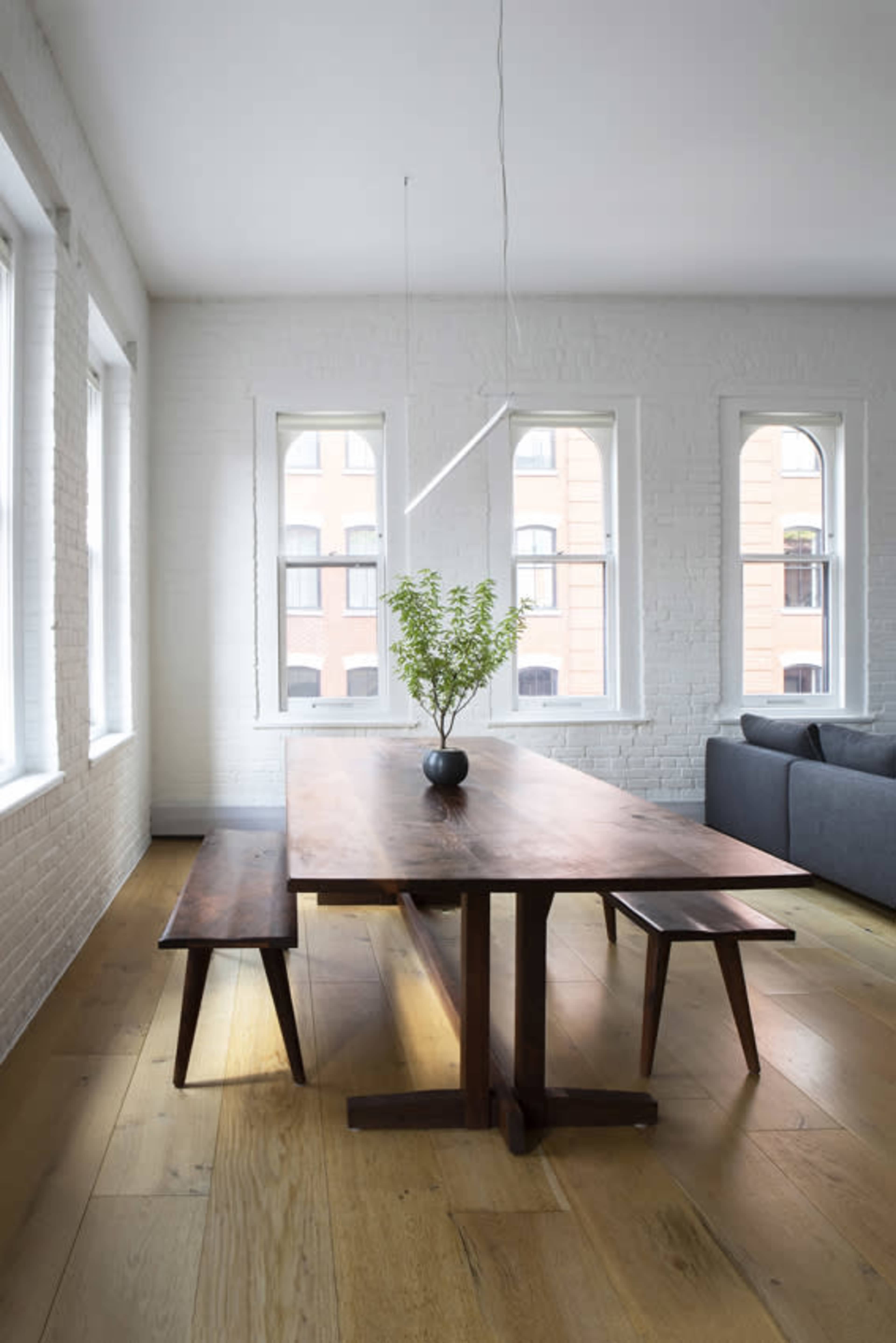 A wooden dining table with a plant centerpiece is positioned between two benches in a bright room with white brick walls and large windows.