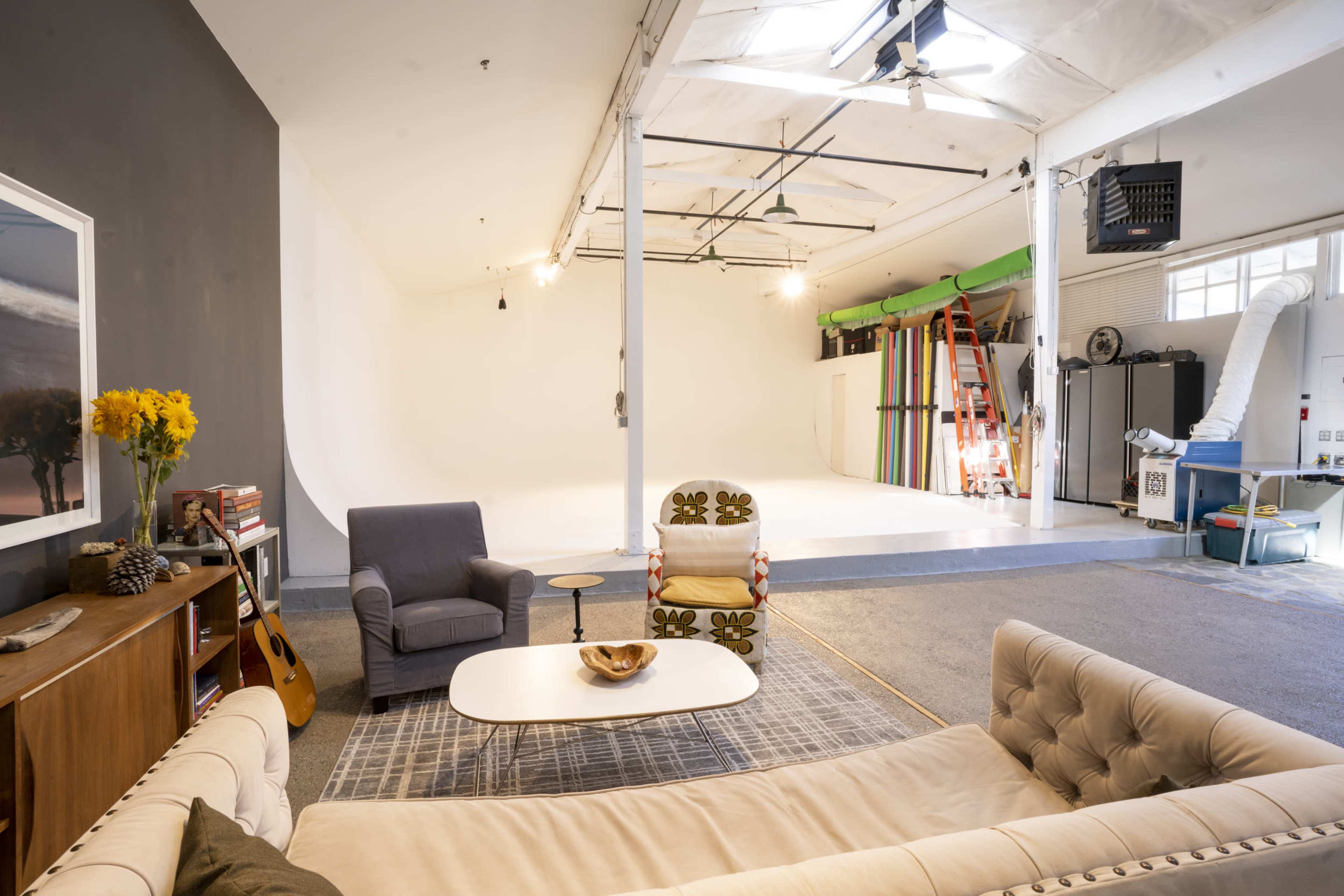 A spacious photo studio features a large white backdrop, a seating area with a sofa and chairs, and various studio equipment along the walls.