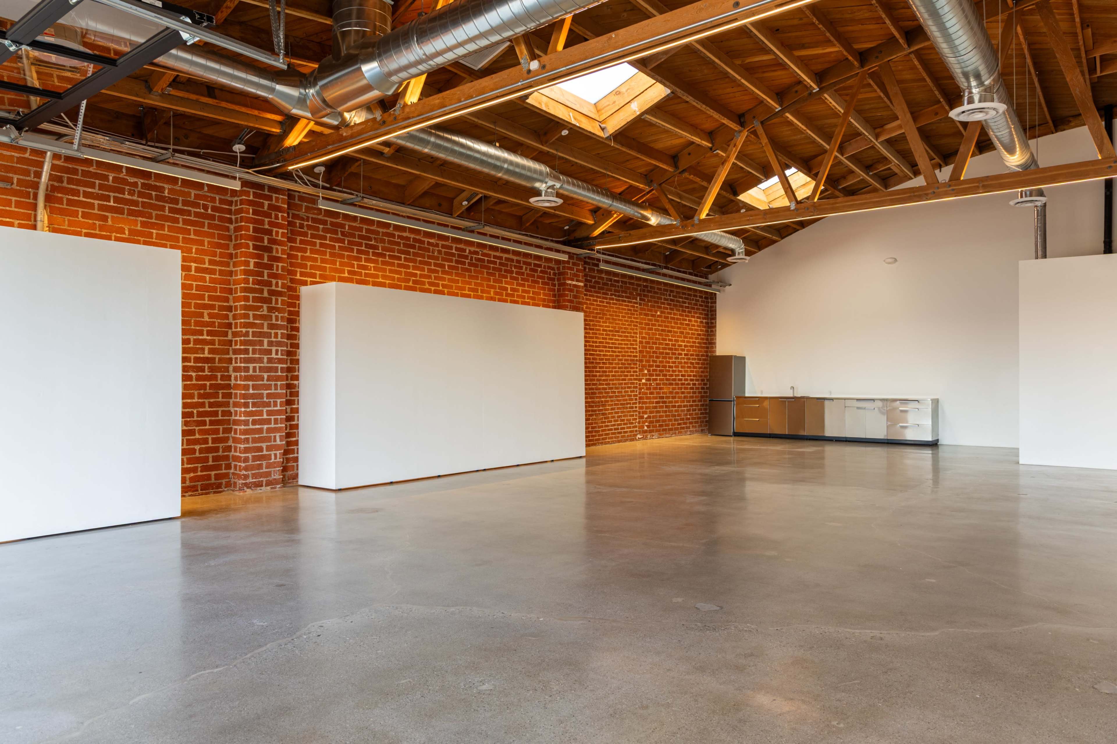The image shows an open, industrial-style space with exposed brick walls, wooden beams, and polished concrete floors, featuring white walls for potential artwork display and a kitchenette area in the corner.