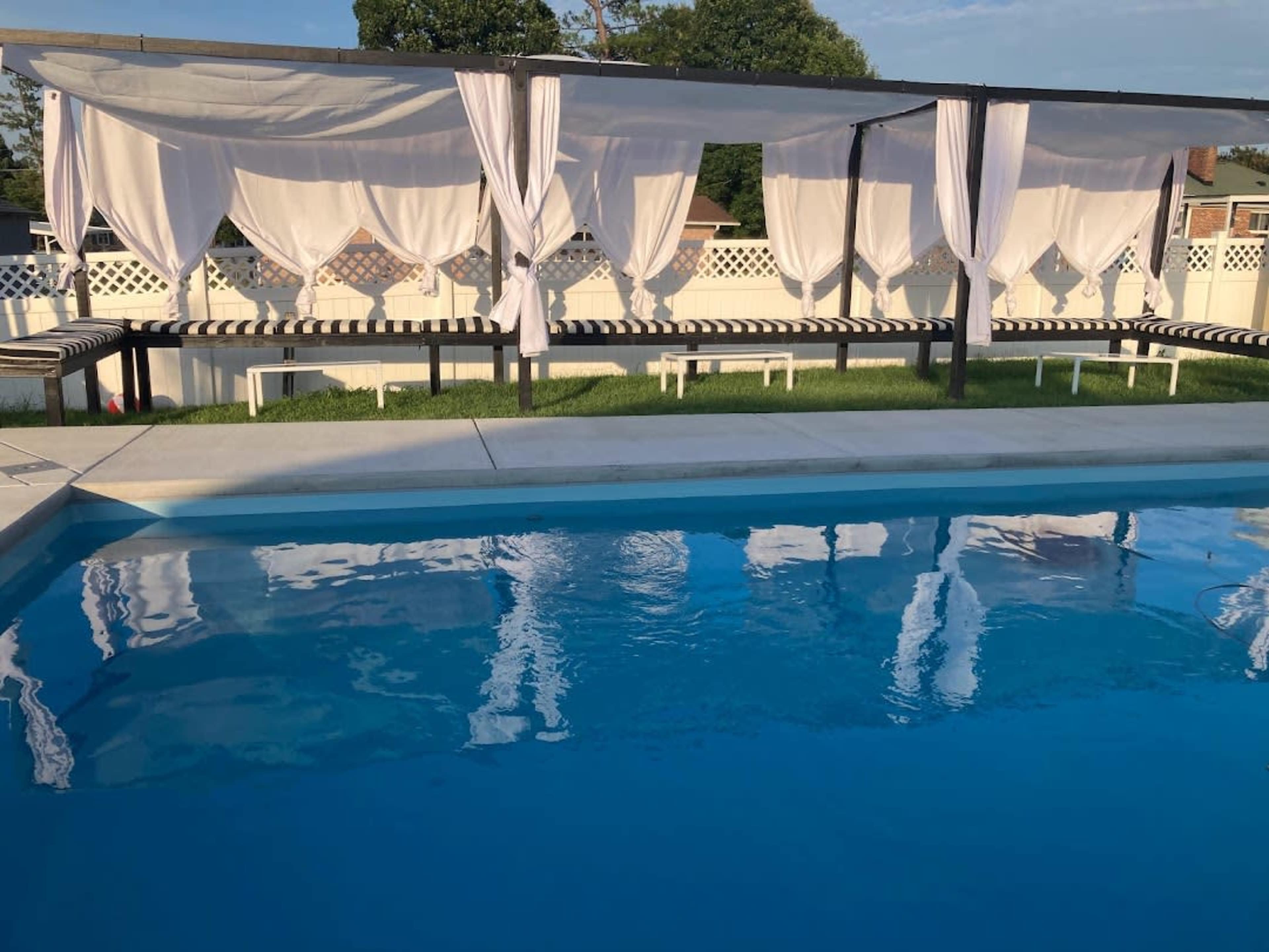 A rectangular swimming pool is surrounded by white, draped cabanas and striped lounge chairs, with lush green grass and a clear blue sky in the background.