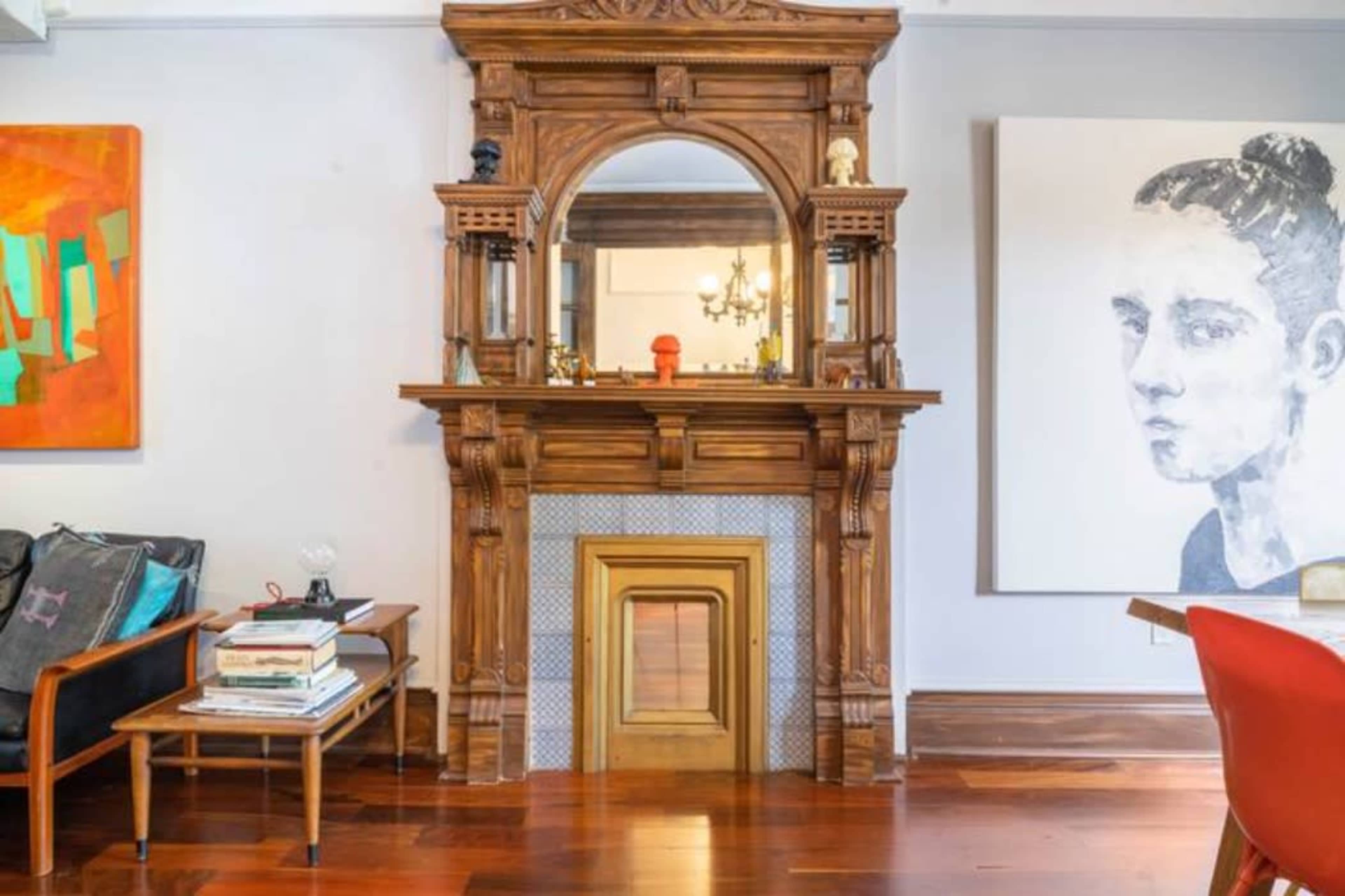 The image shows a wooden fireplace mantel with a large mirror above it, flanked by two artworks on the walls and a chair with magazines in front.