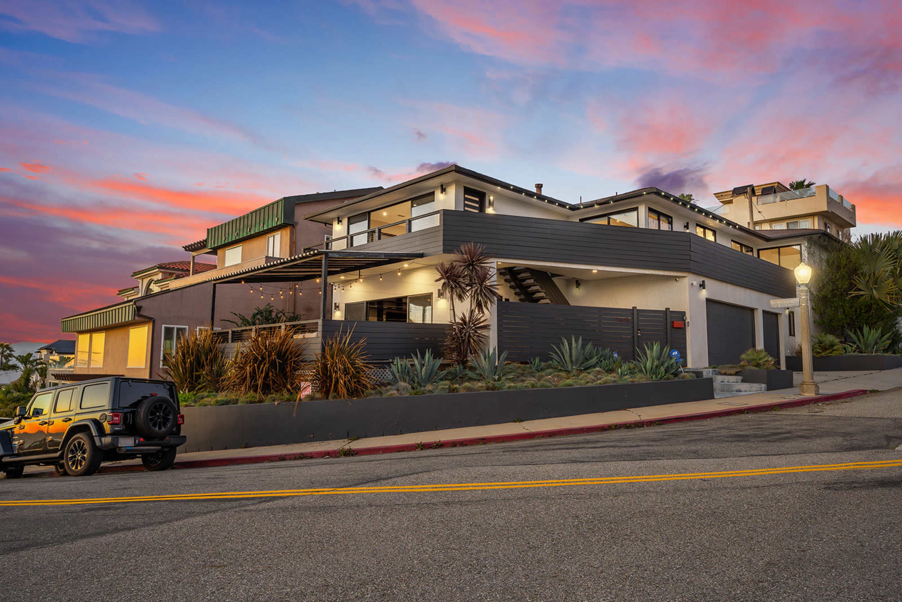 A modern two-story house with a distinct architectural design, surrounded by landscaping and parked vehicles along a sloping street during sunset.