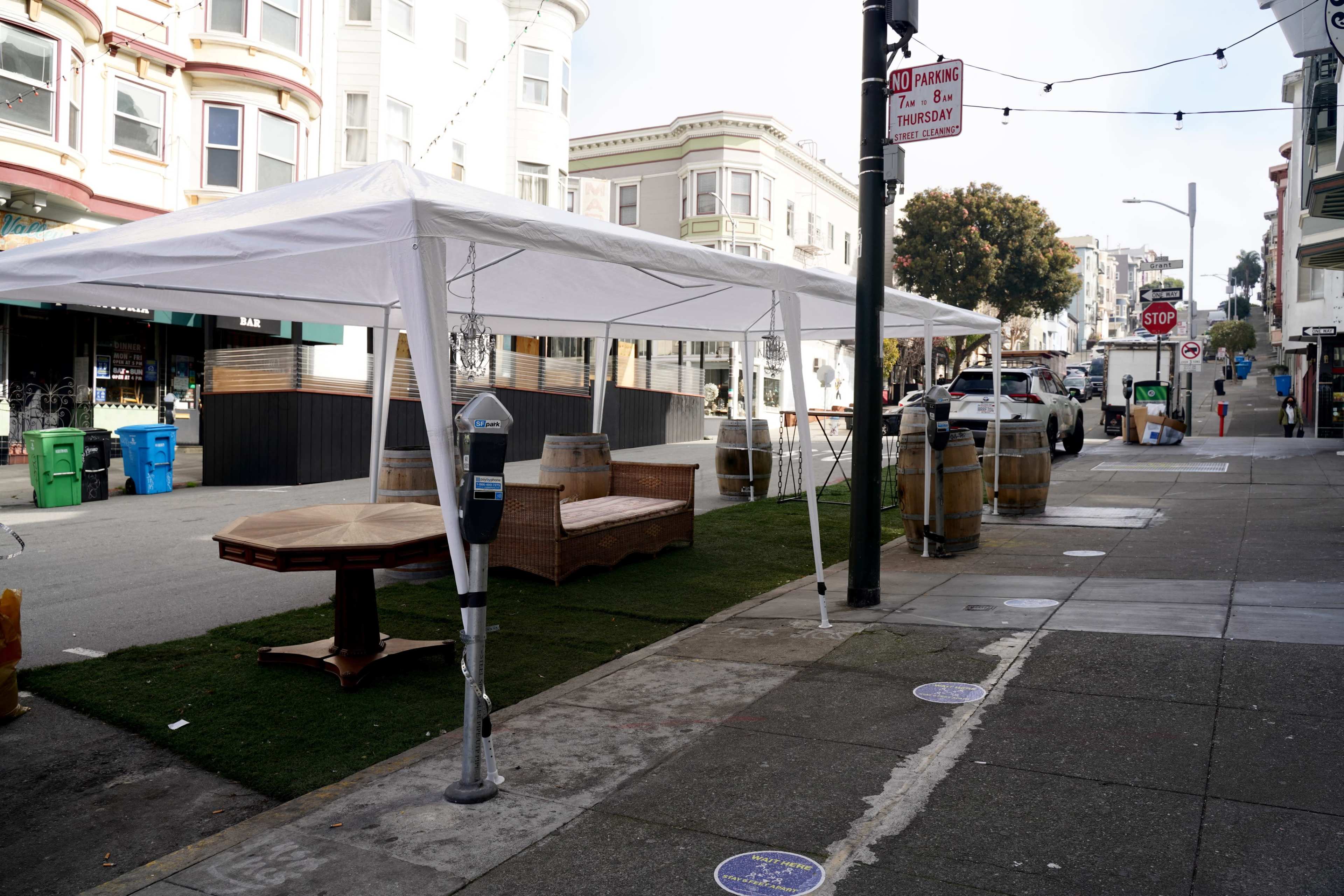 A white tent is set up on a sidewalk next to wooden tables and barrels, with parked cars and trash bins in the background.