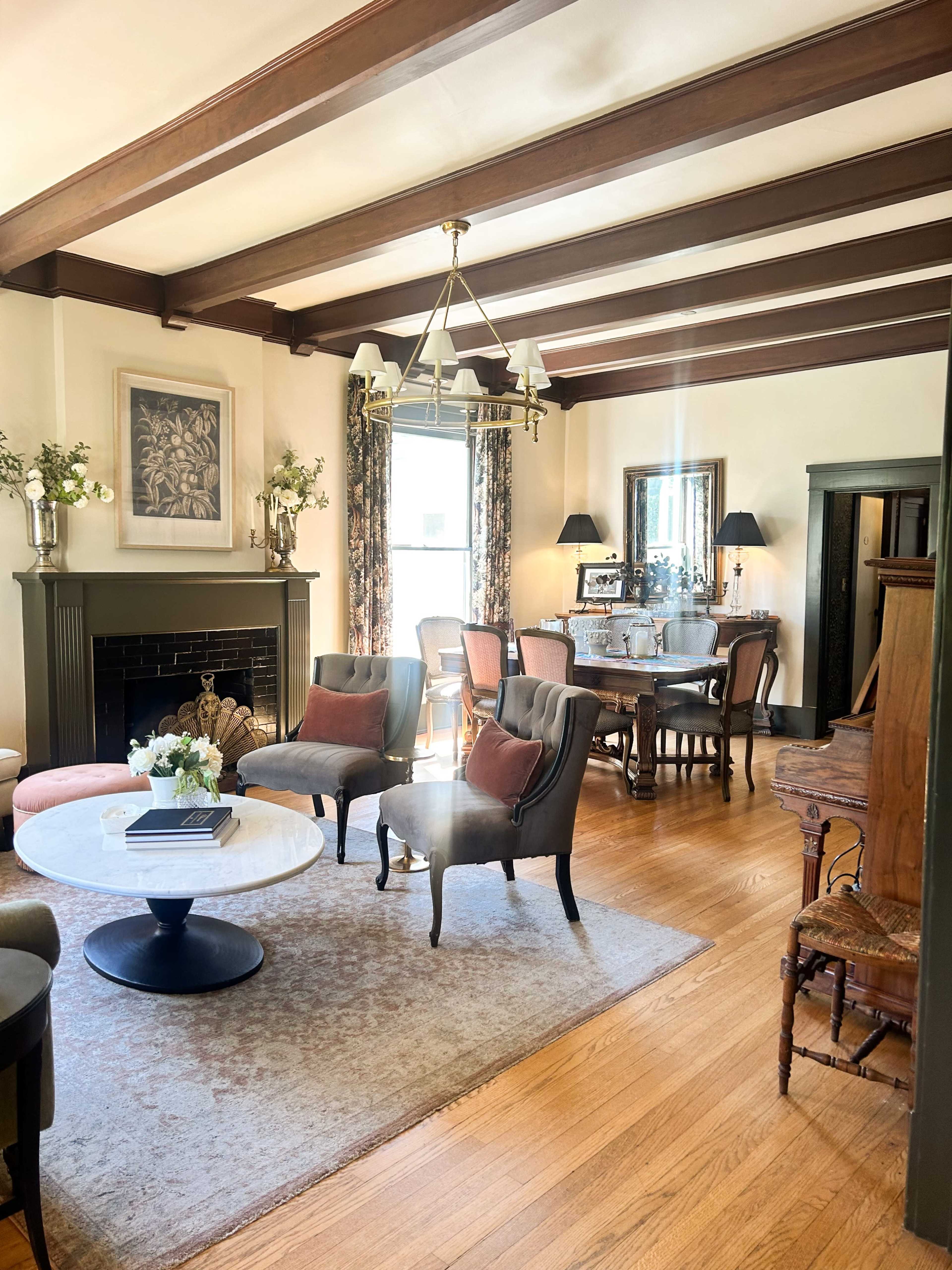 The image shows a cozy living room with wooden beams, a fireplace, and a mixture of seating arrangements, including chairs and a coffee table, leading to a dining area with a table and chairs.