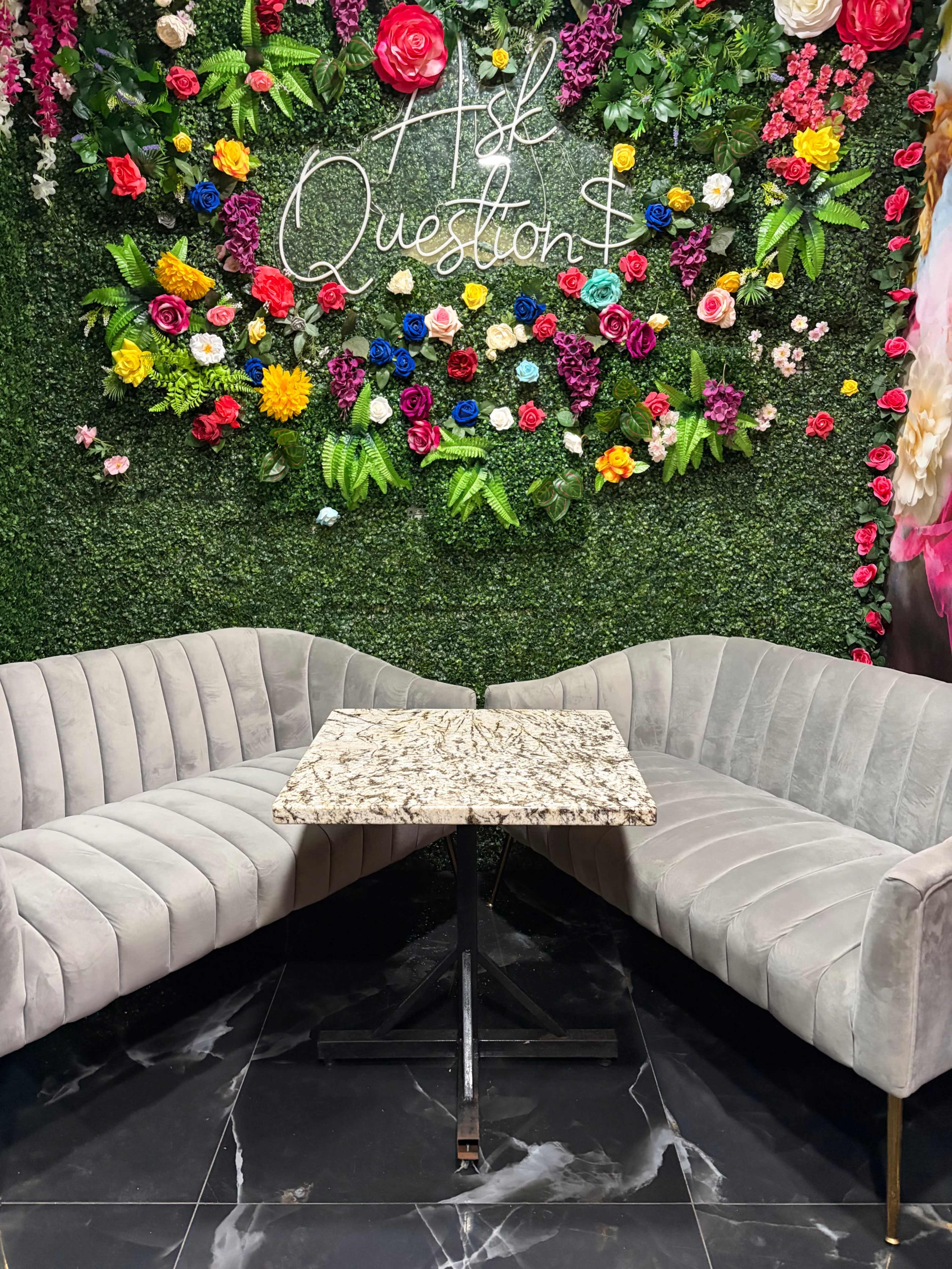 The image shows a cozy seating arrangement with two gray velvet sofas and a decorative wall filled with colorful artificial flowers and greenery.
