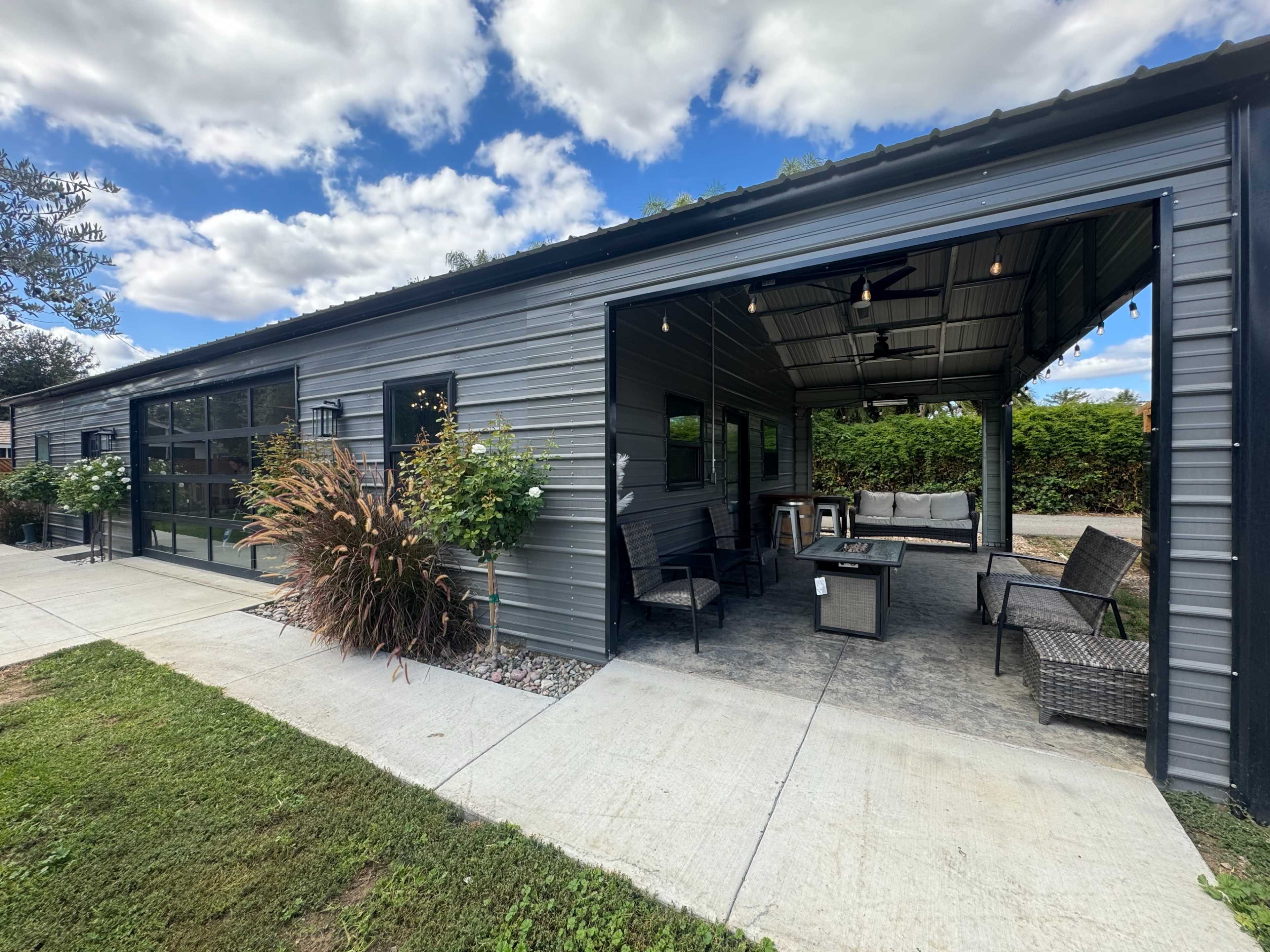 The image shows a modern metal building with a covered patio area that features outdoor seating, surrounded by greenery under a partly cloudy sky.