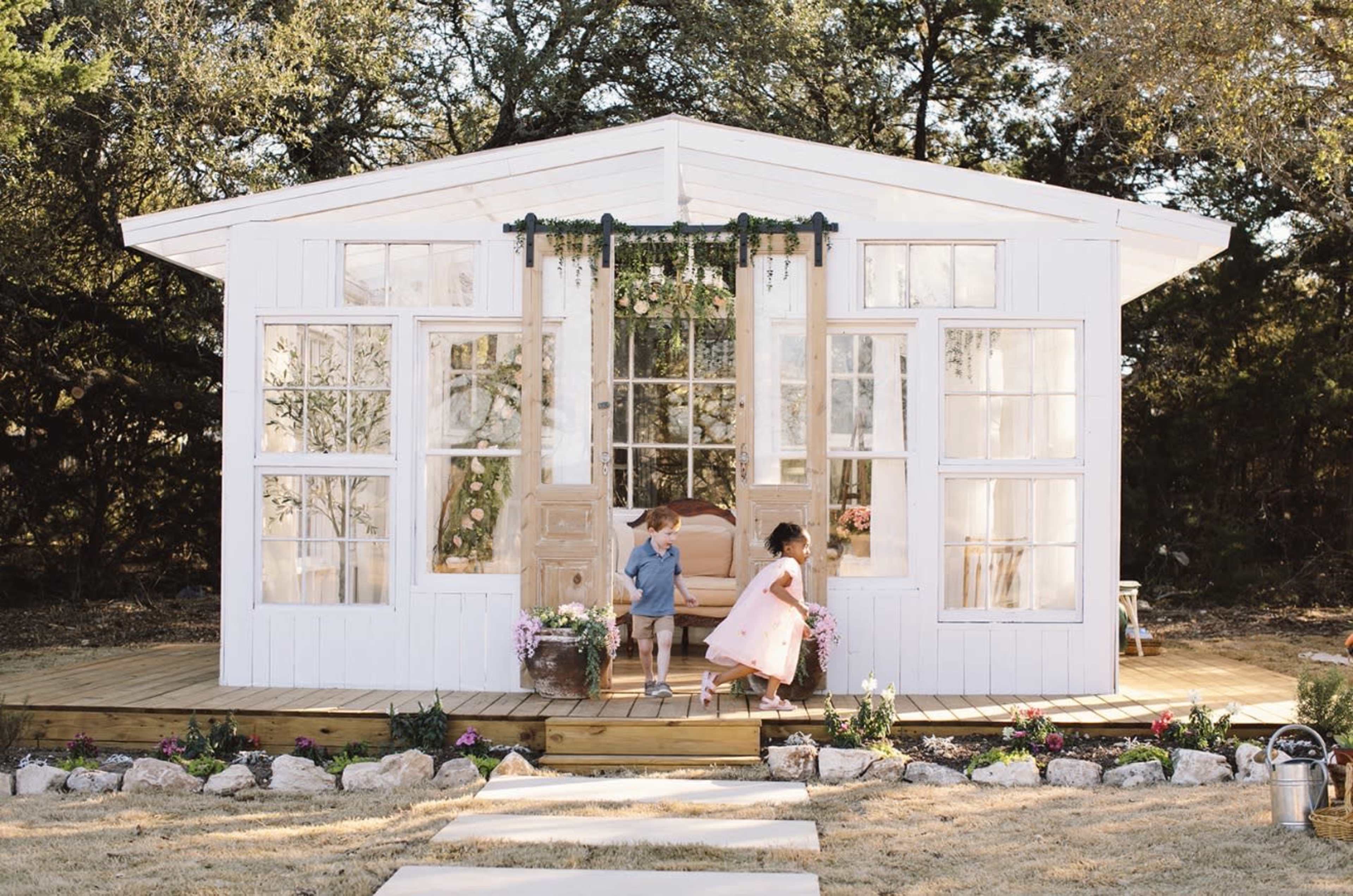 Two children play on the pathway leading to a white, modern-style garden house surrounded by flowers.