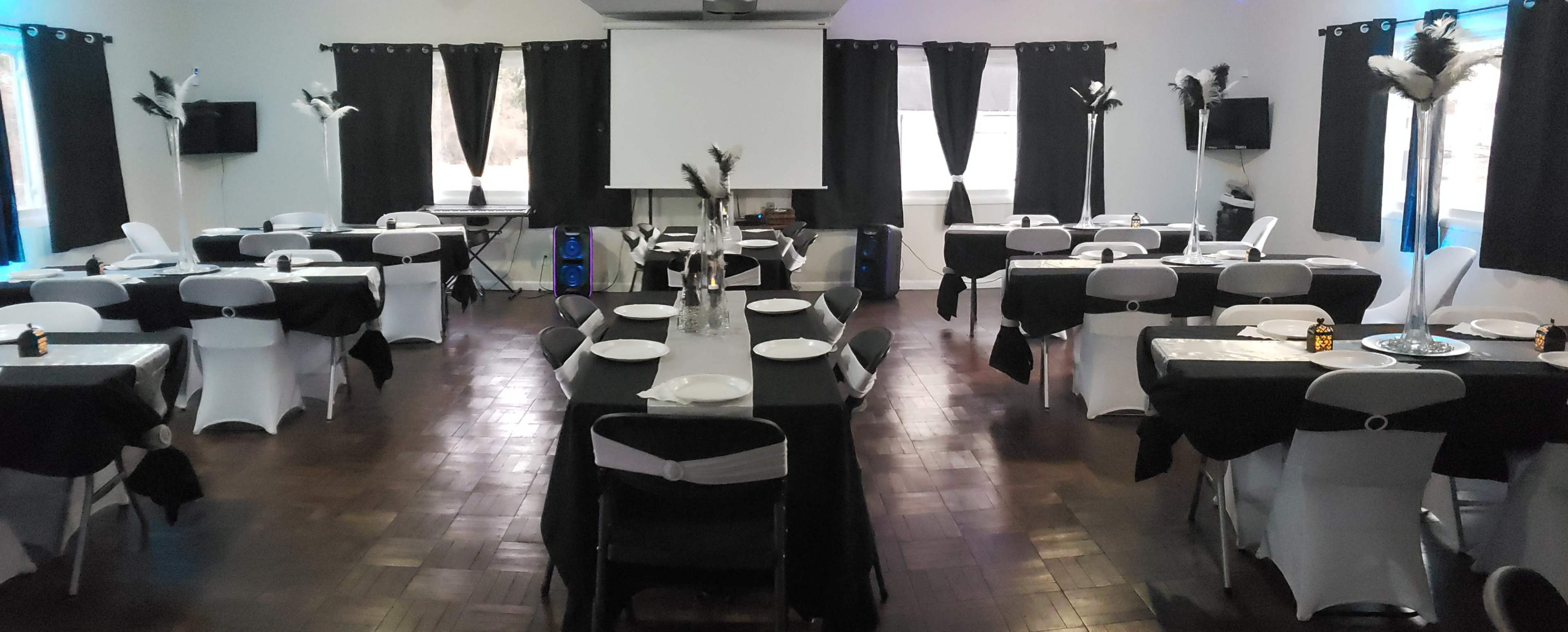 The image shows a dining area set up for an event, featuring tables covered in black tablecloths with white chairs, arranged symmetrically around a central space and decorated with feather centerpieces.
