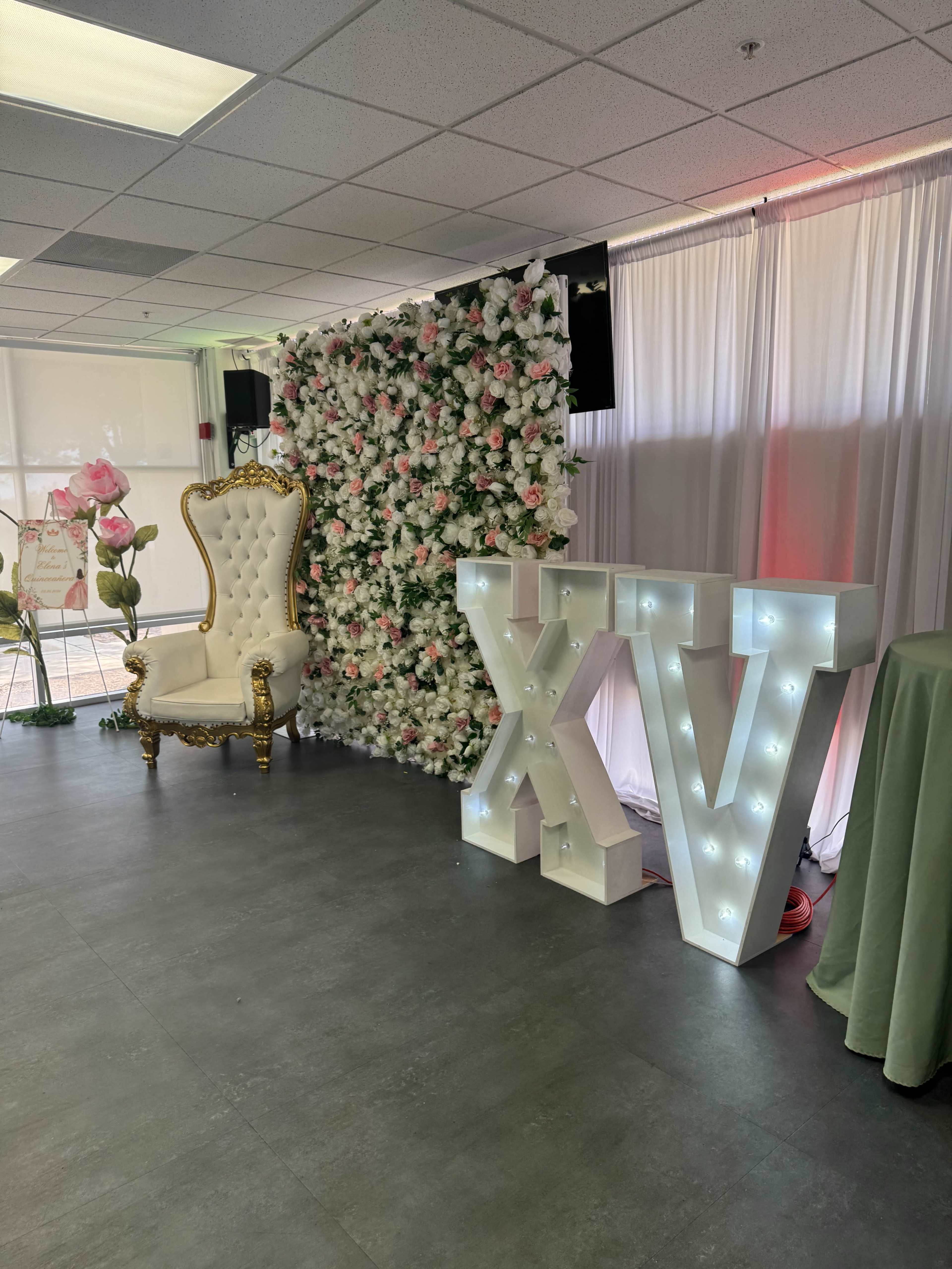 The image shows a decorated event space featuring a tall, white floral backdrop with pink flowers, a gold-accented throne chair to the left, and illuminated letters "XV" arranged prominently on the floor.