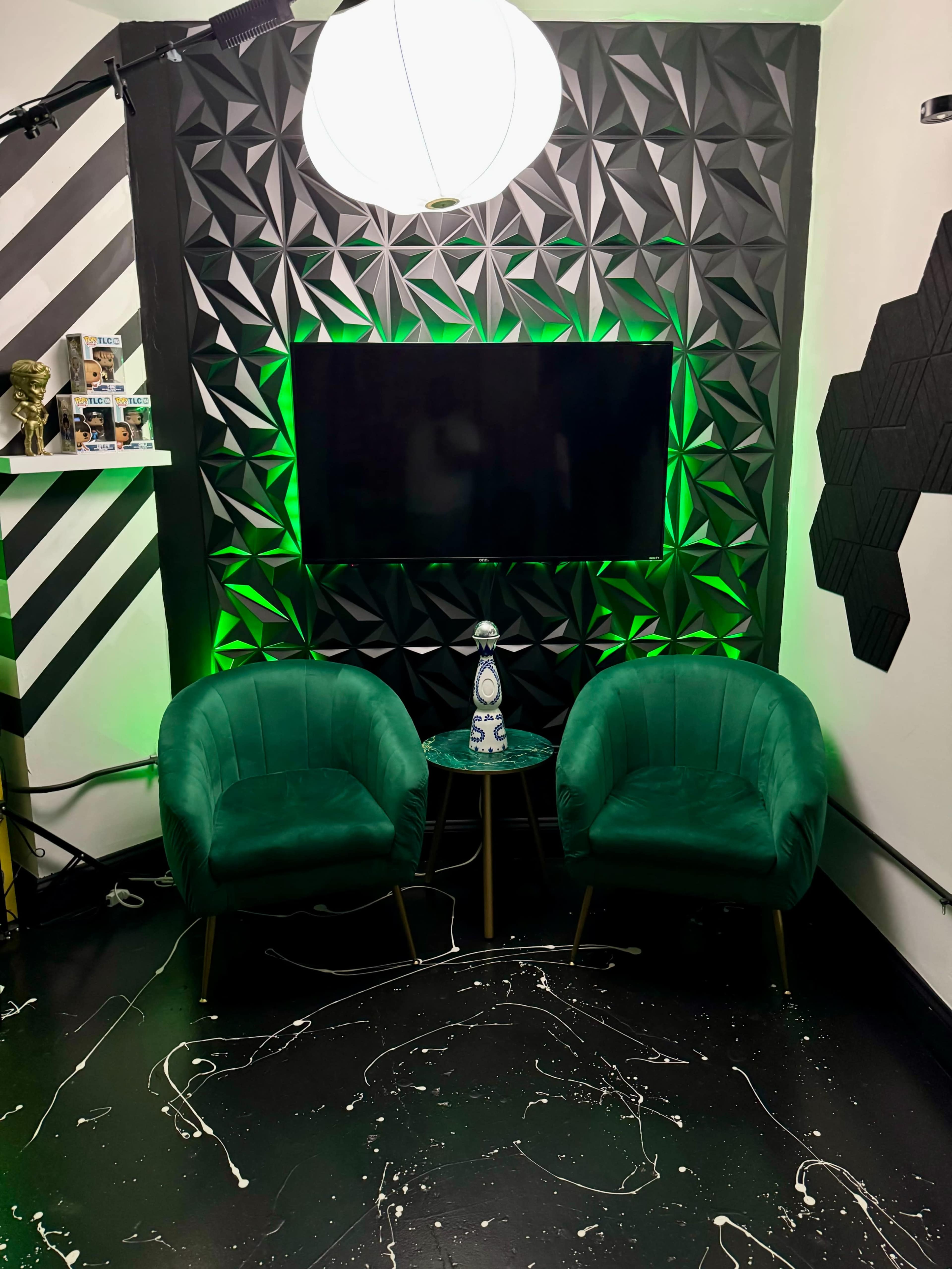 The scene features two green chairs facing a mounted television, surrounded by a textured black wall with green lighting and a patterned floor.
