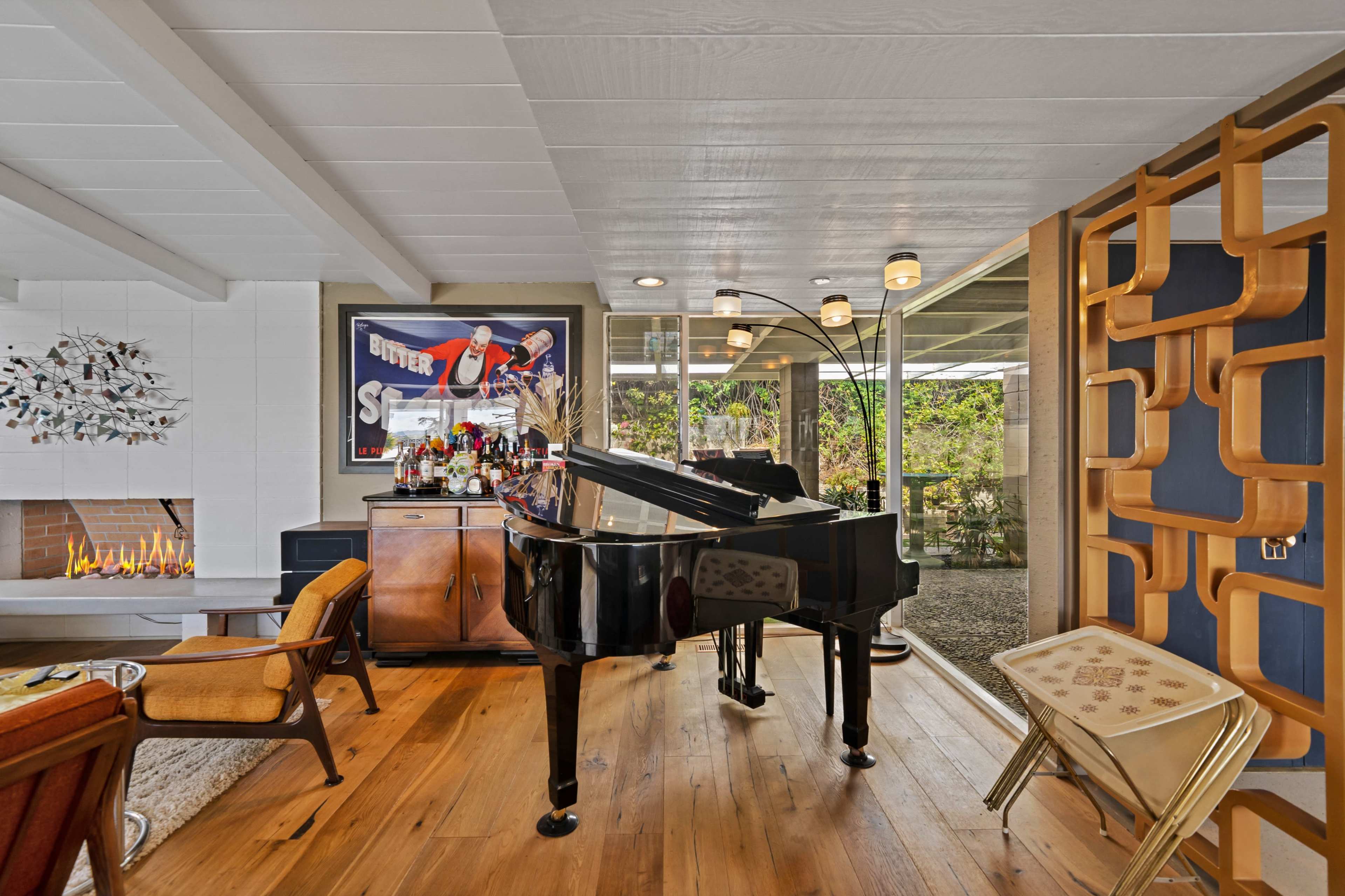 A grand piano is positioned near a modern fireplace, with a bar setup and contemporary furniture in a well-lit living area that opens to a garden view.