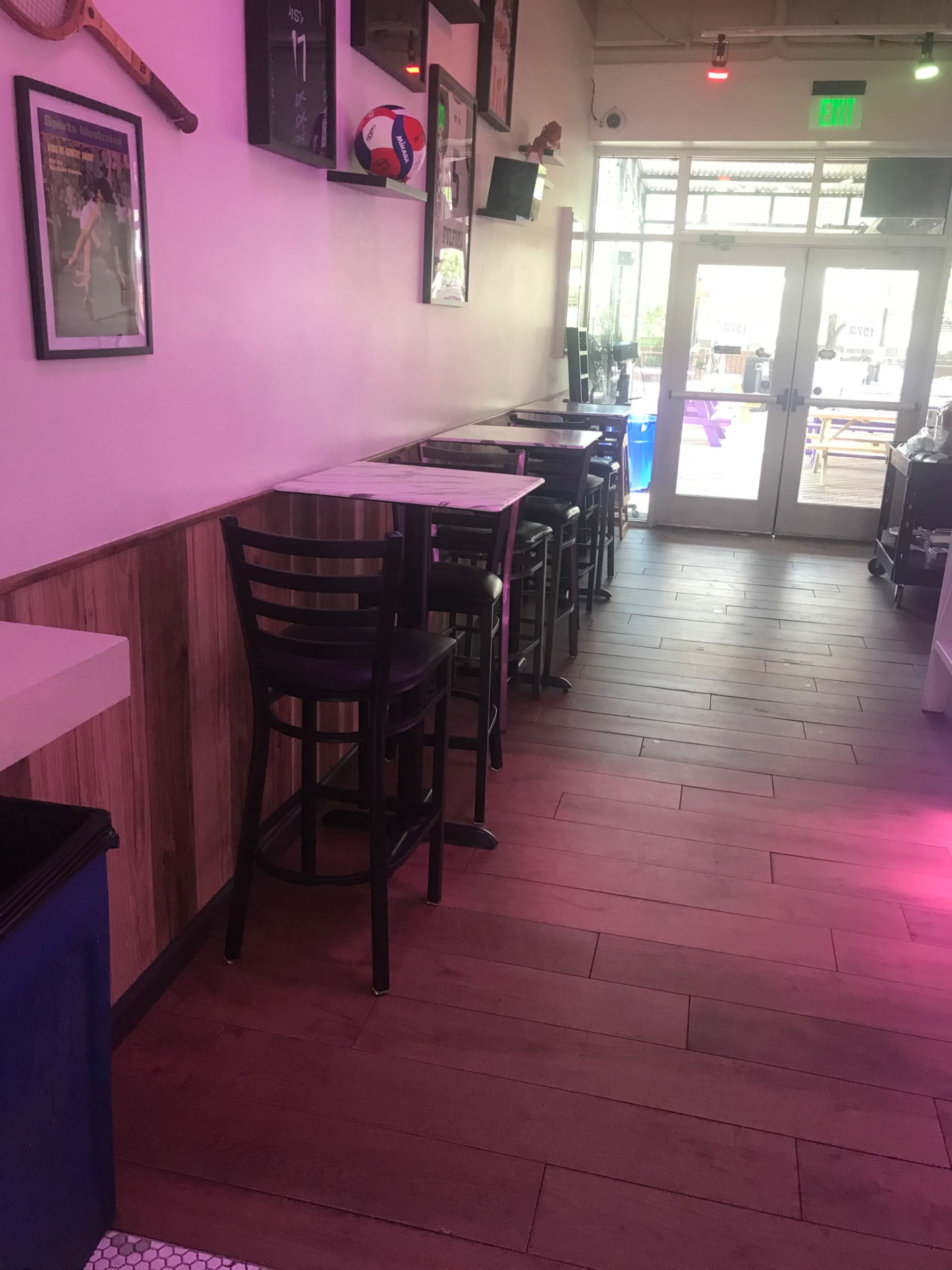 The image shows a narrow corridor in a restaurant with bar-height tables and chairs along one side, leading to large glass doors.