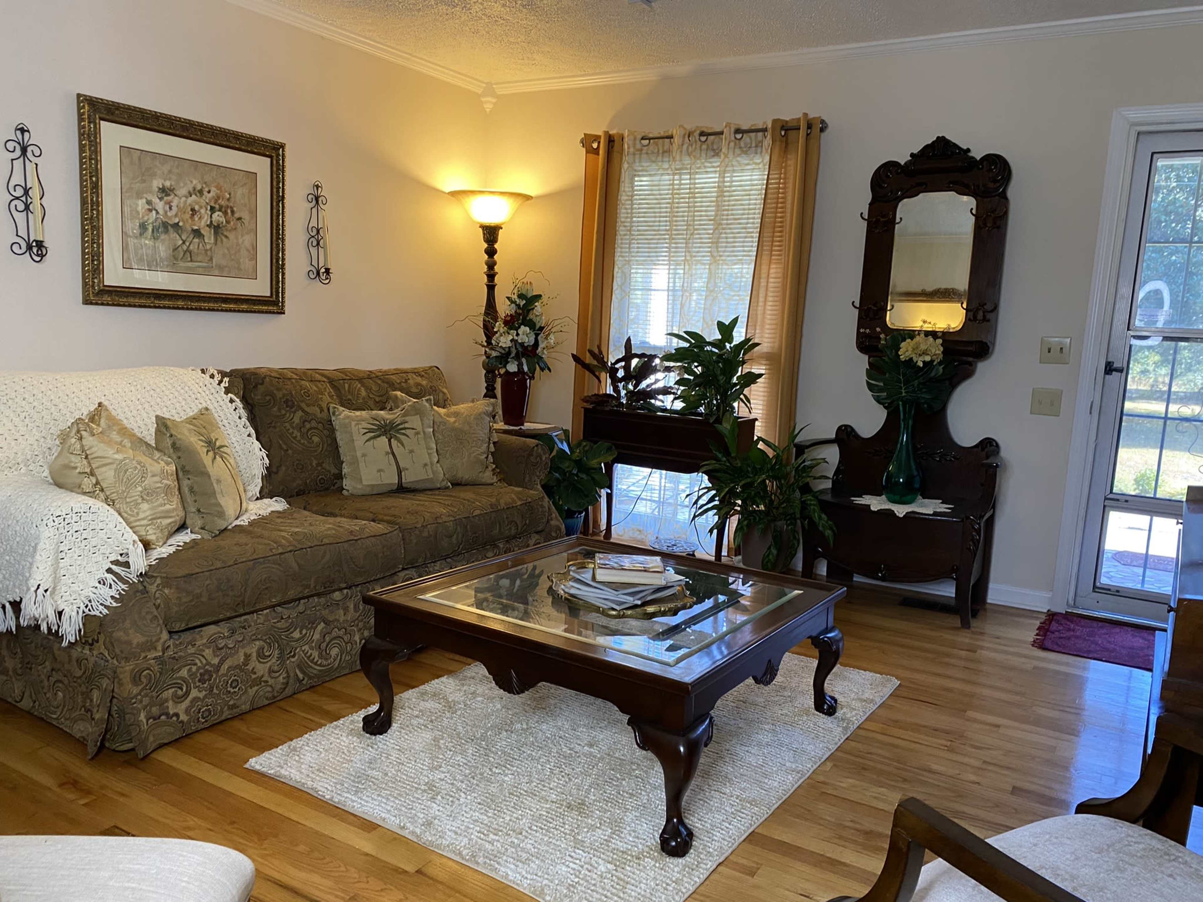 A cozy living room features a brown sofa with decorative pillows, a glass coffee table, houseplants, and a wall mirror, illuminated by a floor lamp.