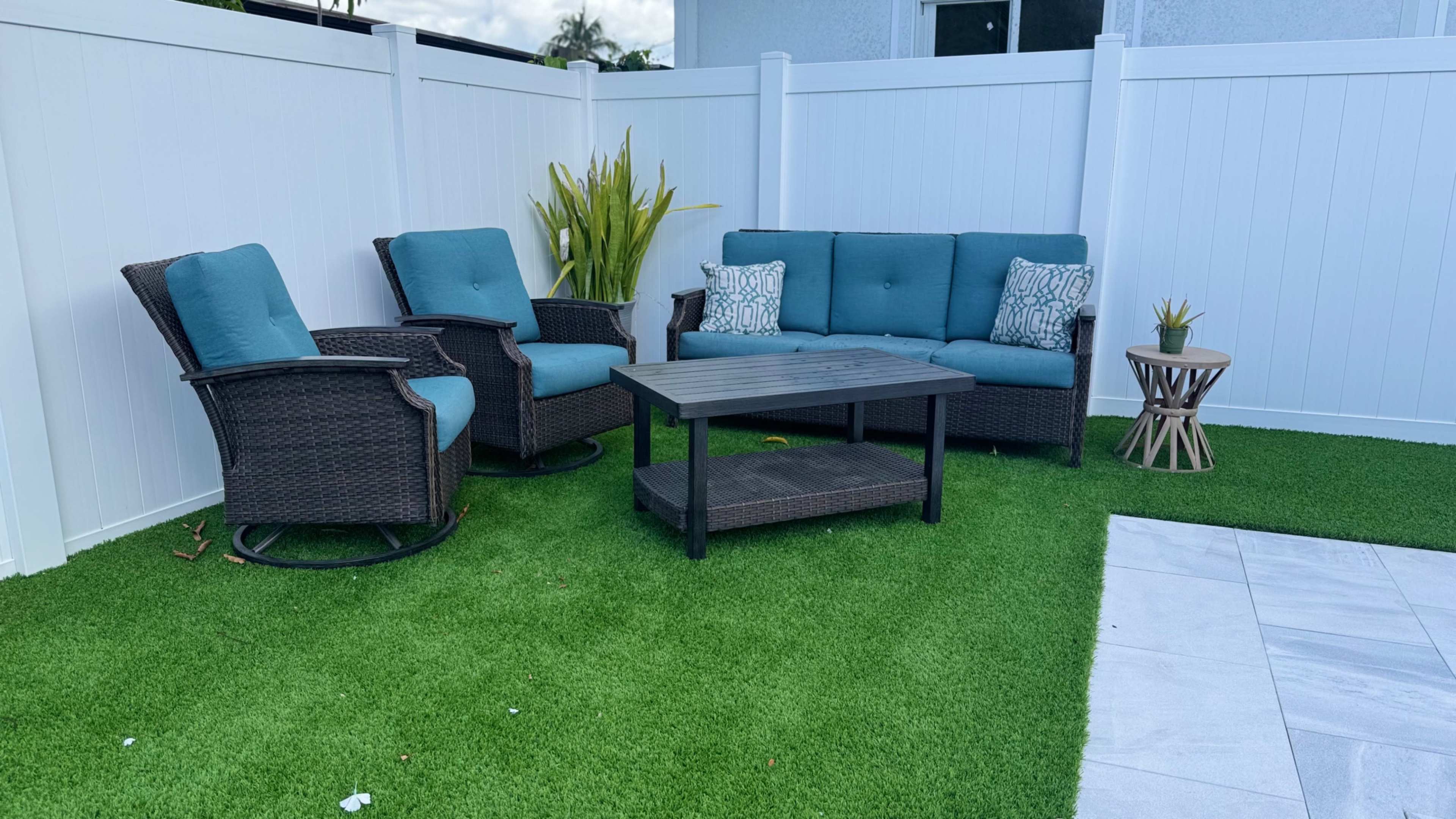 The image shows a seating area with a sofa and two chairs arranged around a coffee table on artificial grass, surrounded by white fences and a decorative plant.