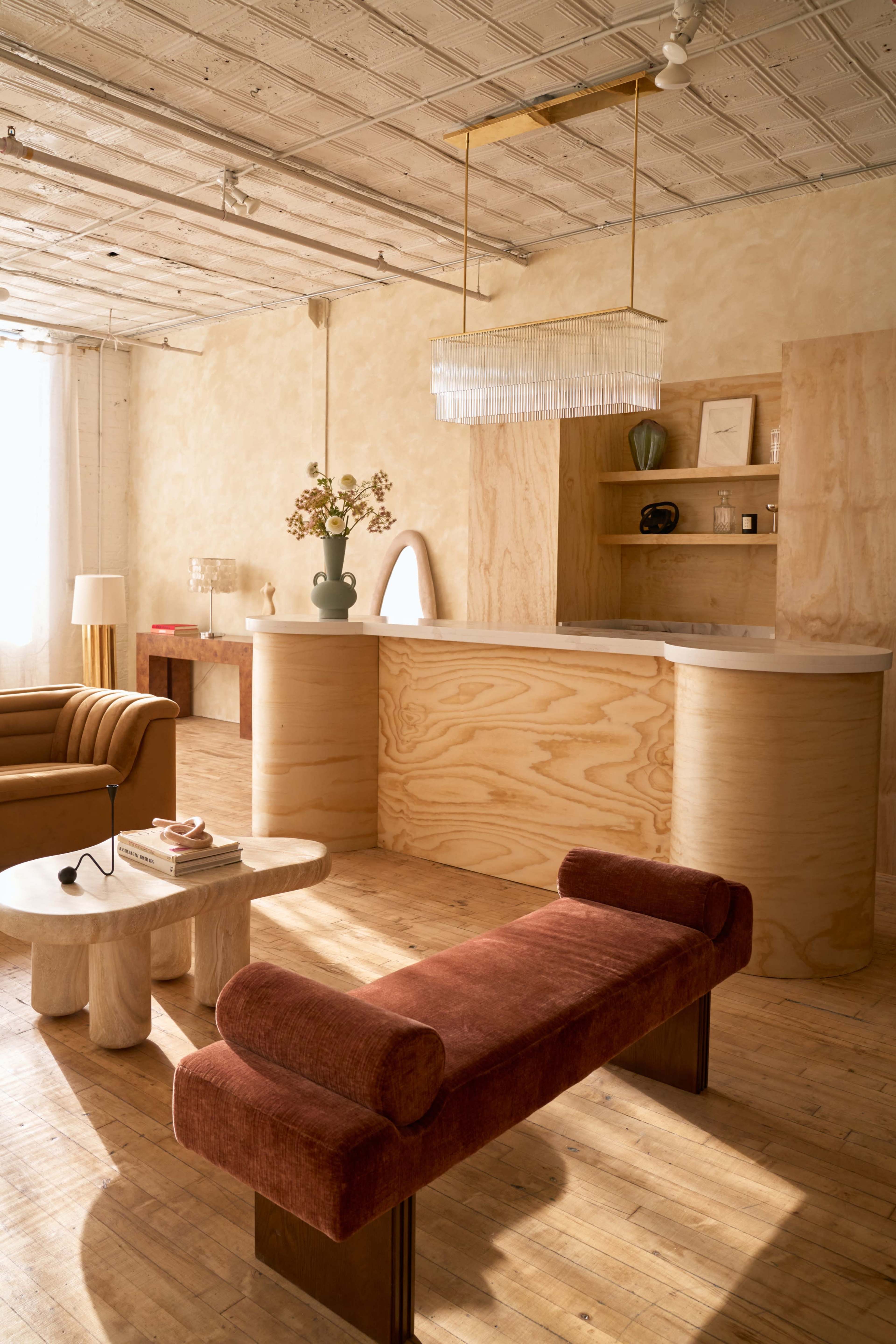 The image features a modern interior space with wood-paneled walls, a kitchen island made of plywood, and a seating area that includes a curved sofa, a round coffee table, and a plush bench.