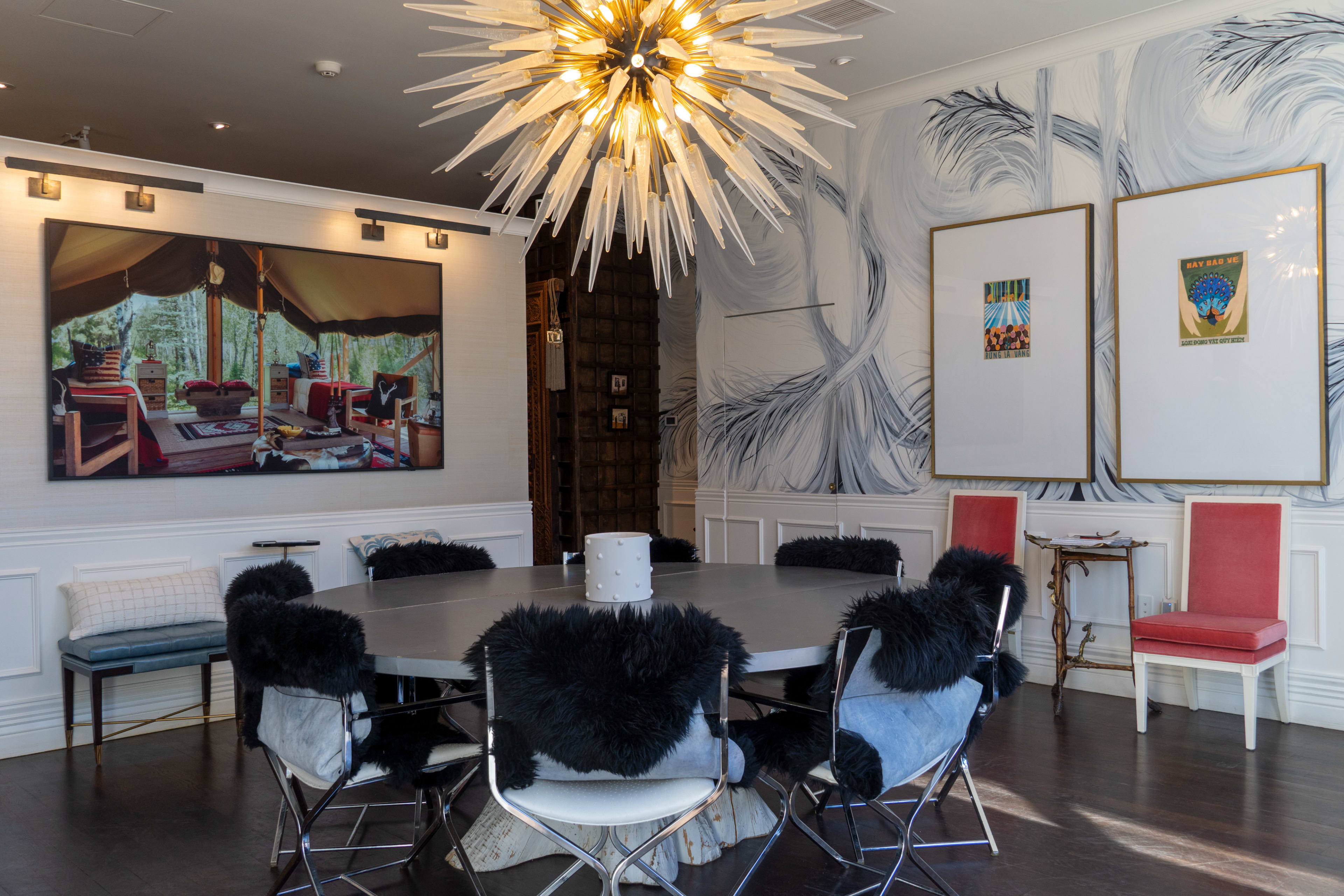 The image shows a modern dining room with a circular table surrounded by chairs with black fur accents, illuminated by a starburst chandelier, and adorned with a large wall art piece and two framed posters.