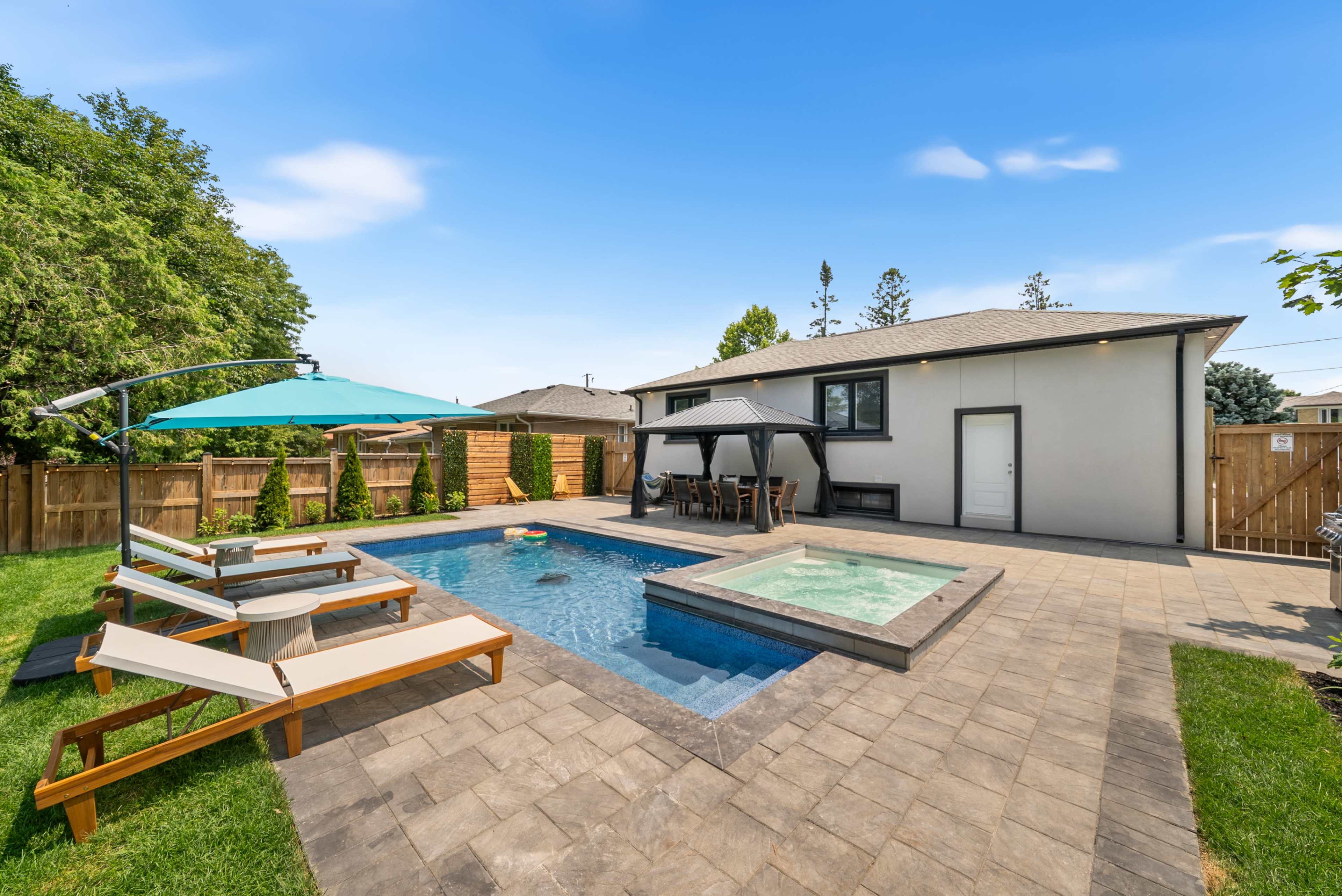 The image shows a residential backyard featuring a swimming pool, a hot tub, lounge chairs, and a covered dining area.