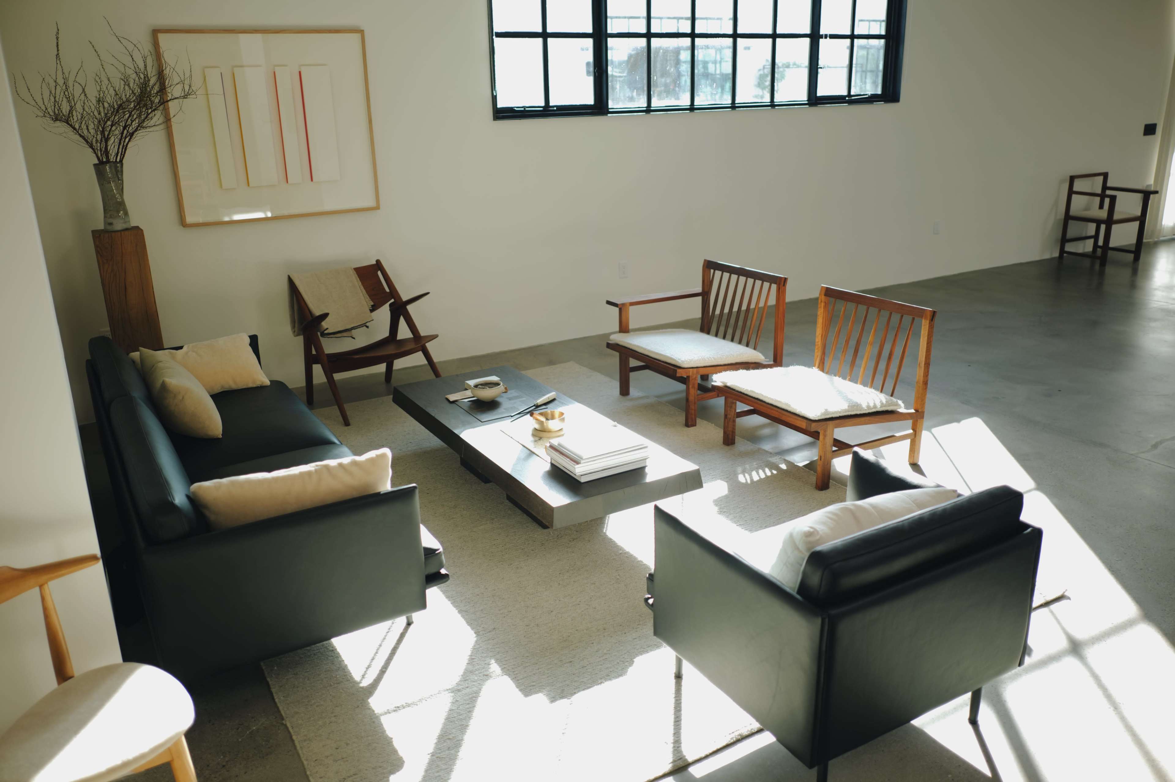 The image shows a minimalist living room with two black sofas, several wooden chairs, a low coffee table, and large windows that allow natural light to fill the space.