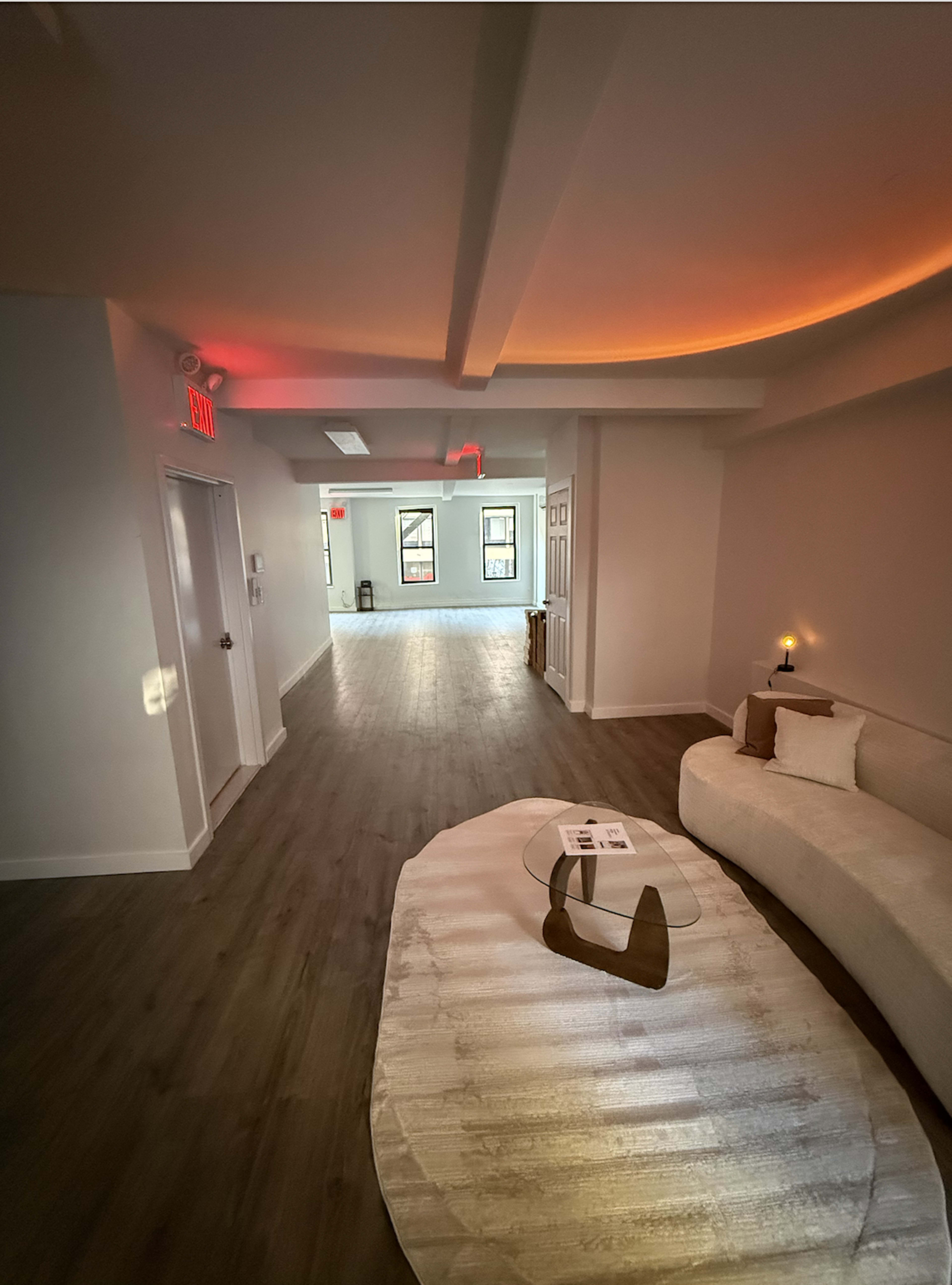 The image shows a spacious, well-lit hallway leading to a room with large windows, alongside a minimalist seating area with a curved couch and a small table.