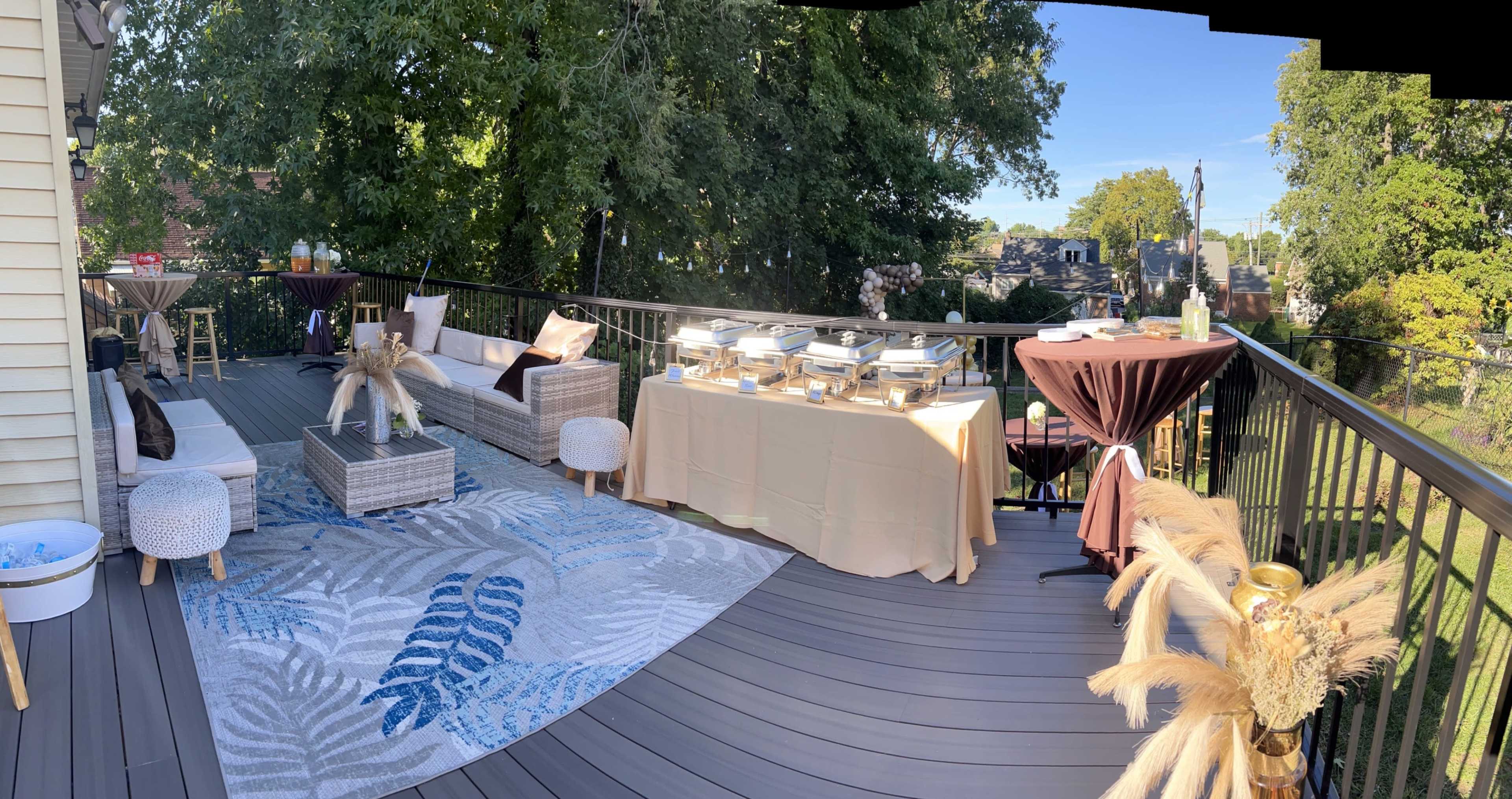 A spacious outdoor deck is set up for a gathering, featuring a buffet table with food trays, seating areas, and decorative accents.
