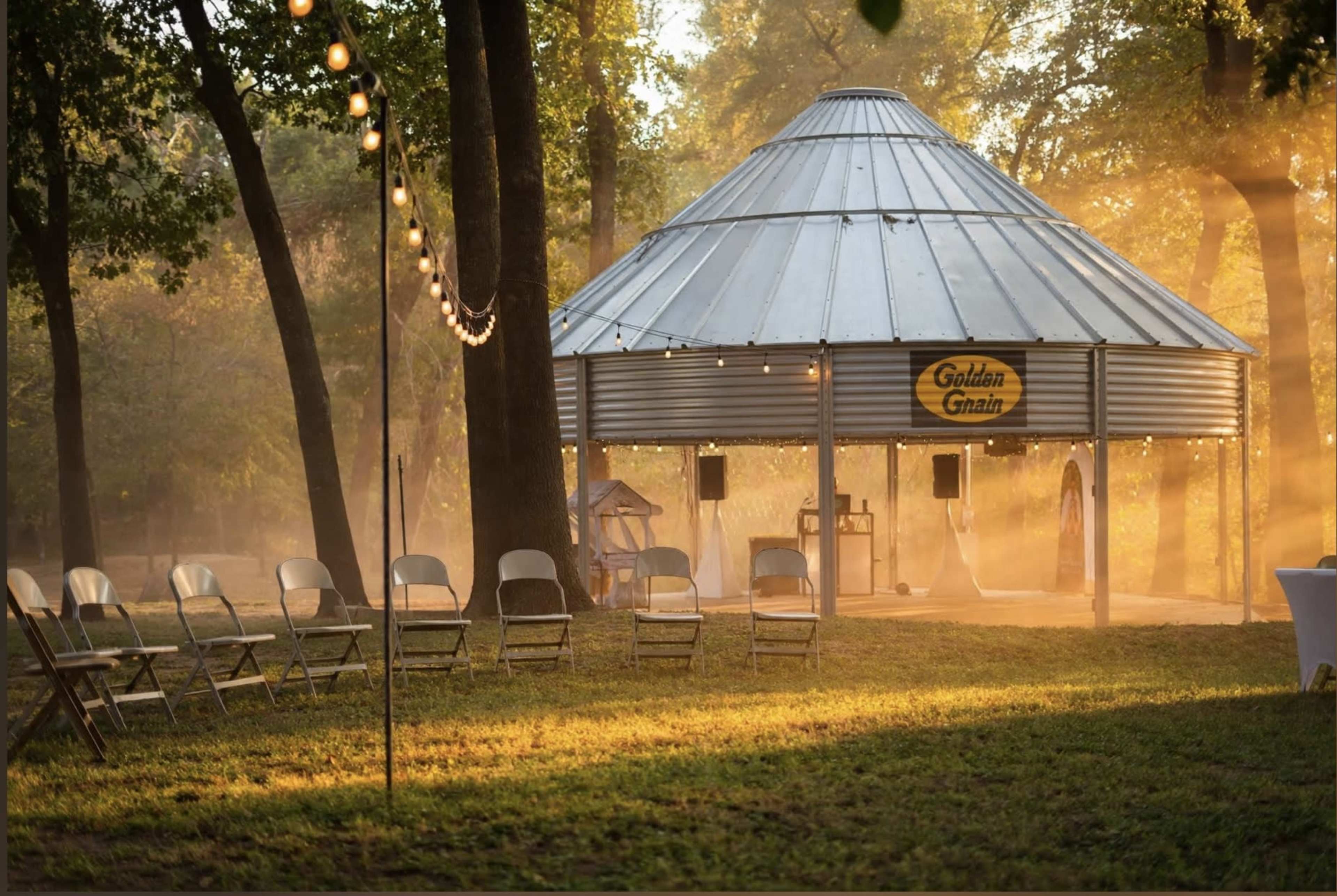 A circular pavilion with a metal roof is surrounded by chairs and illuminated by golden sunlight filtering through the trees in a serene outdoor setting.