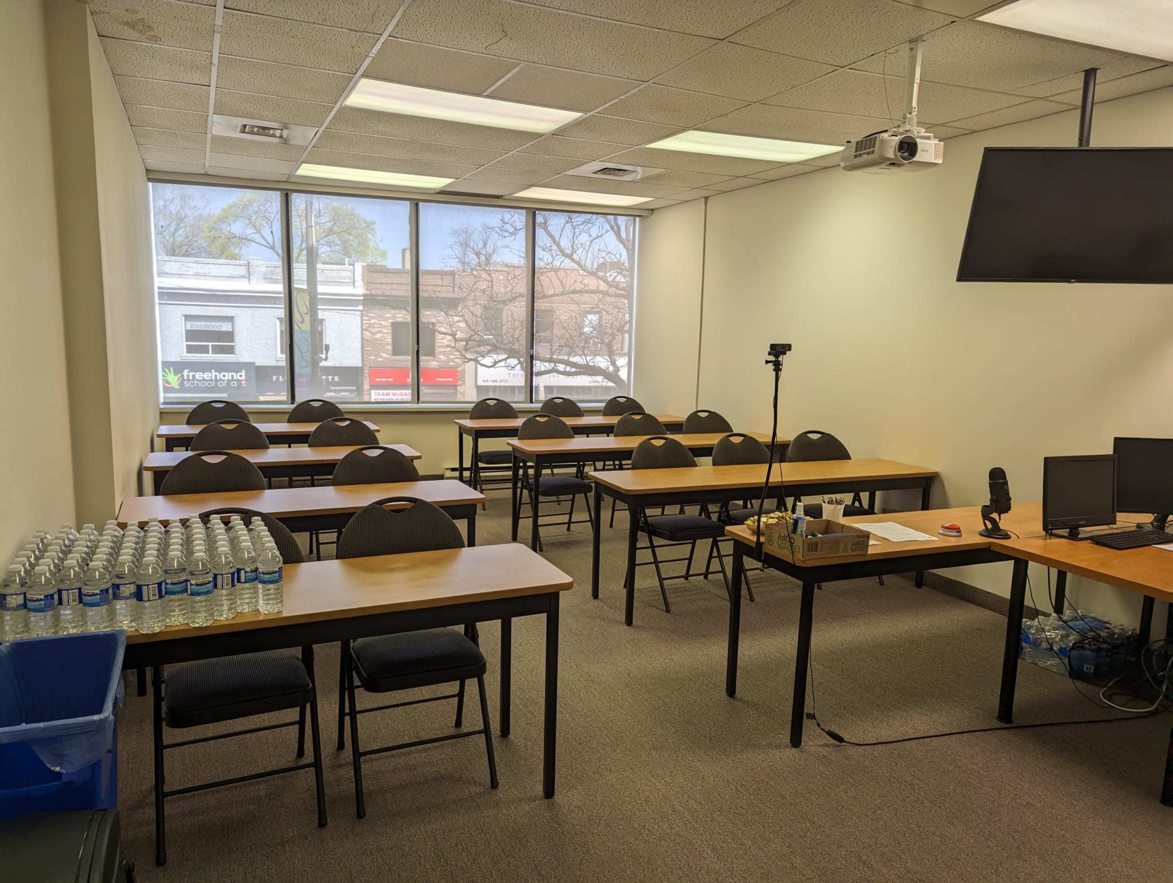 A classroom is set up with rows of tables and chairs, equipped with a projector and a monitor, alongside a wall of windows displaying a view of the street outside.