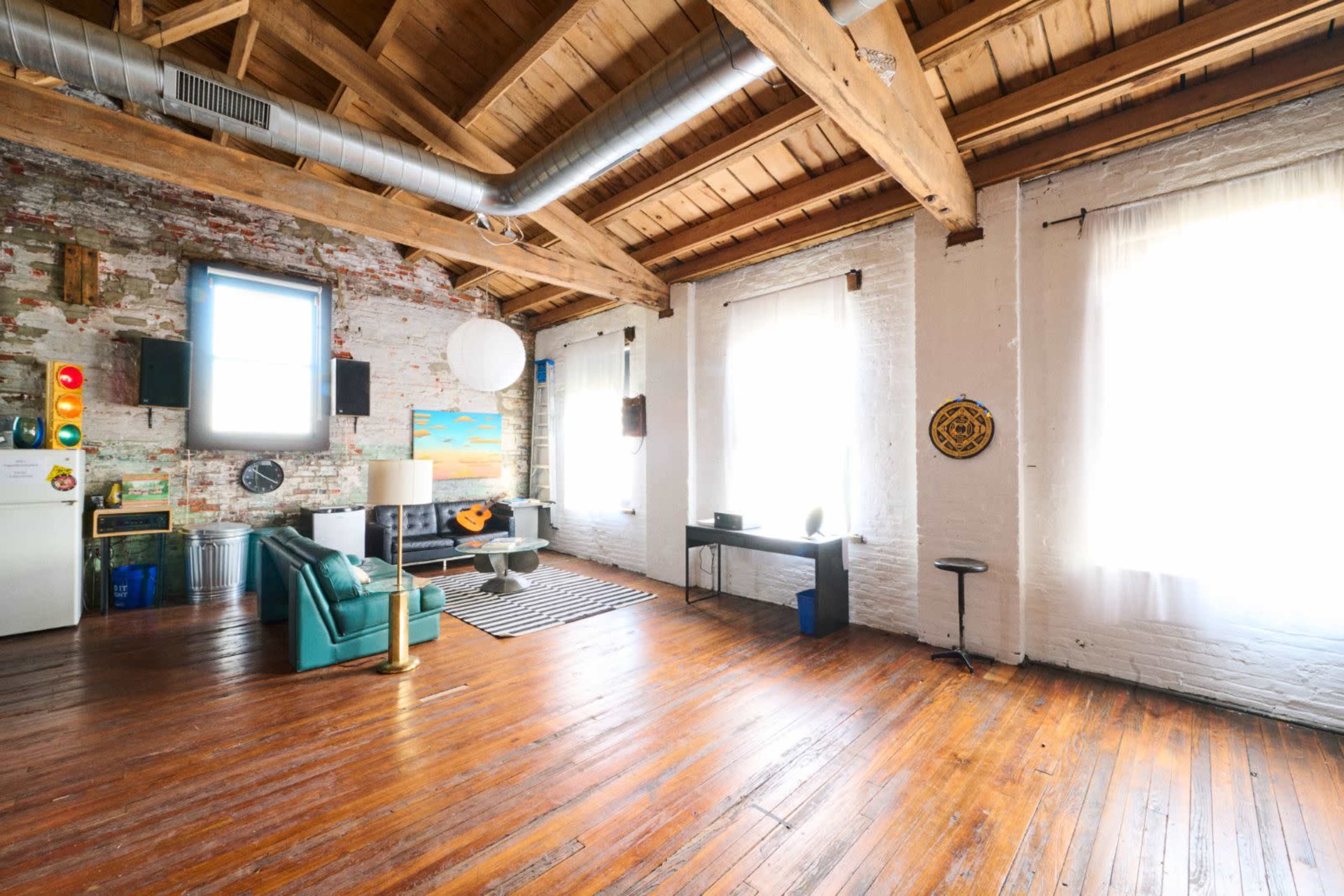 A spacious, industrial-style room with wooden beams, large windows, and a mix of furniture, including a turquoise couch and a small table.