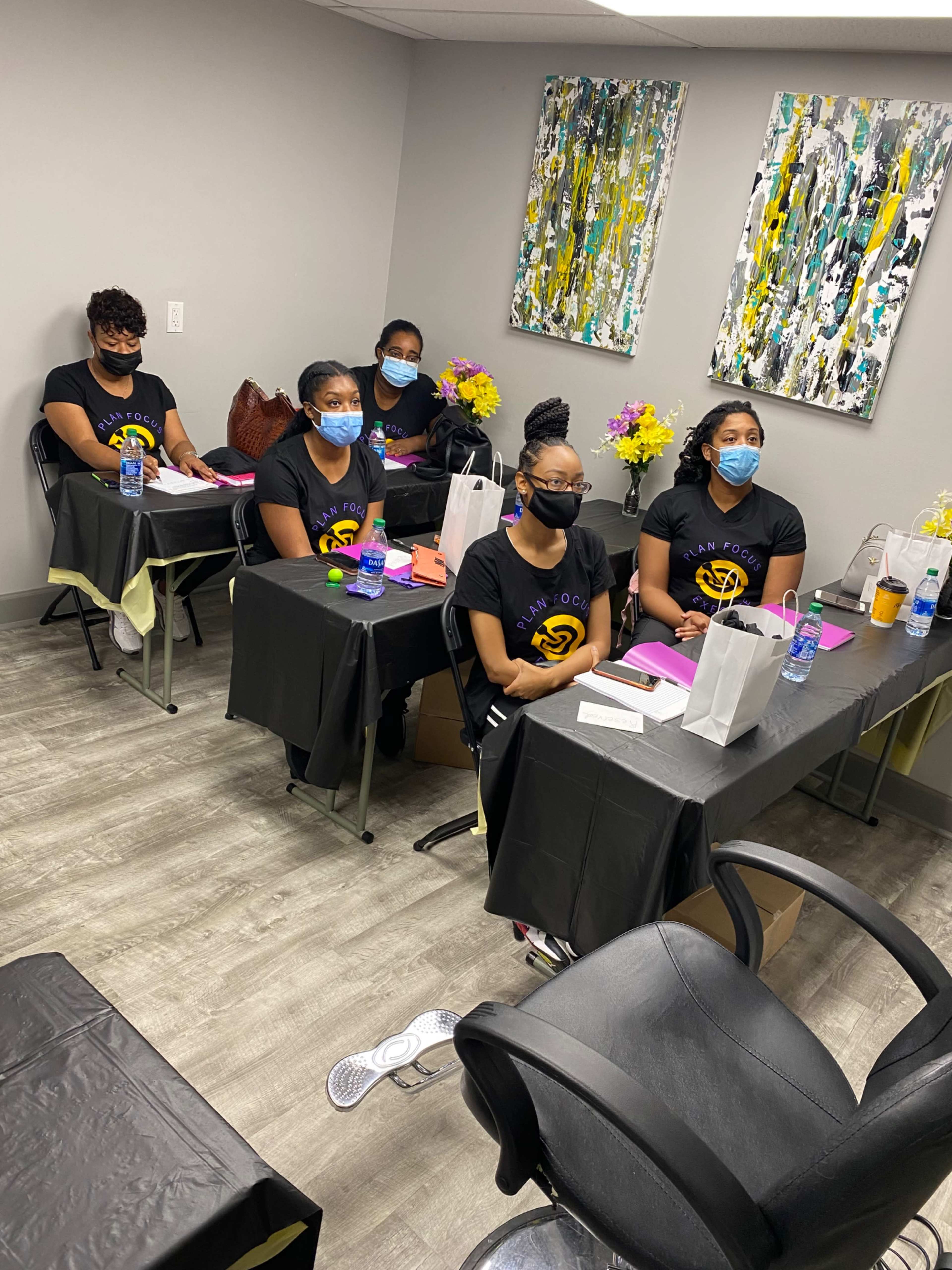 A group of six individuals wearing black shirts and face masks sit at tables in a room decorated with paintings and flower arrangements.