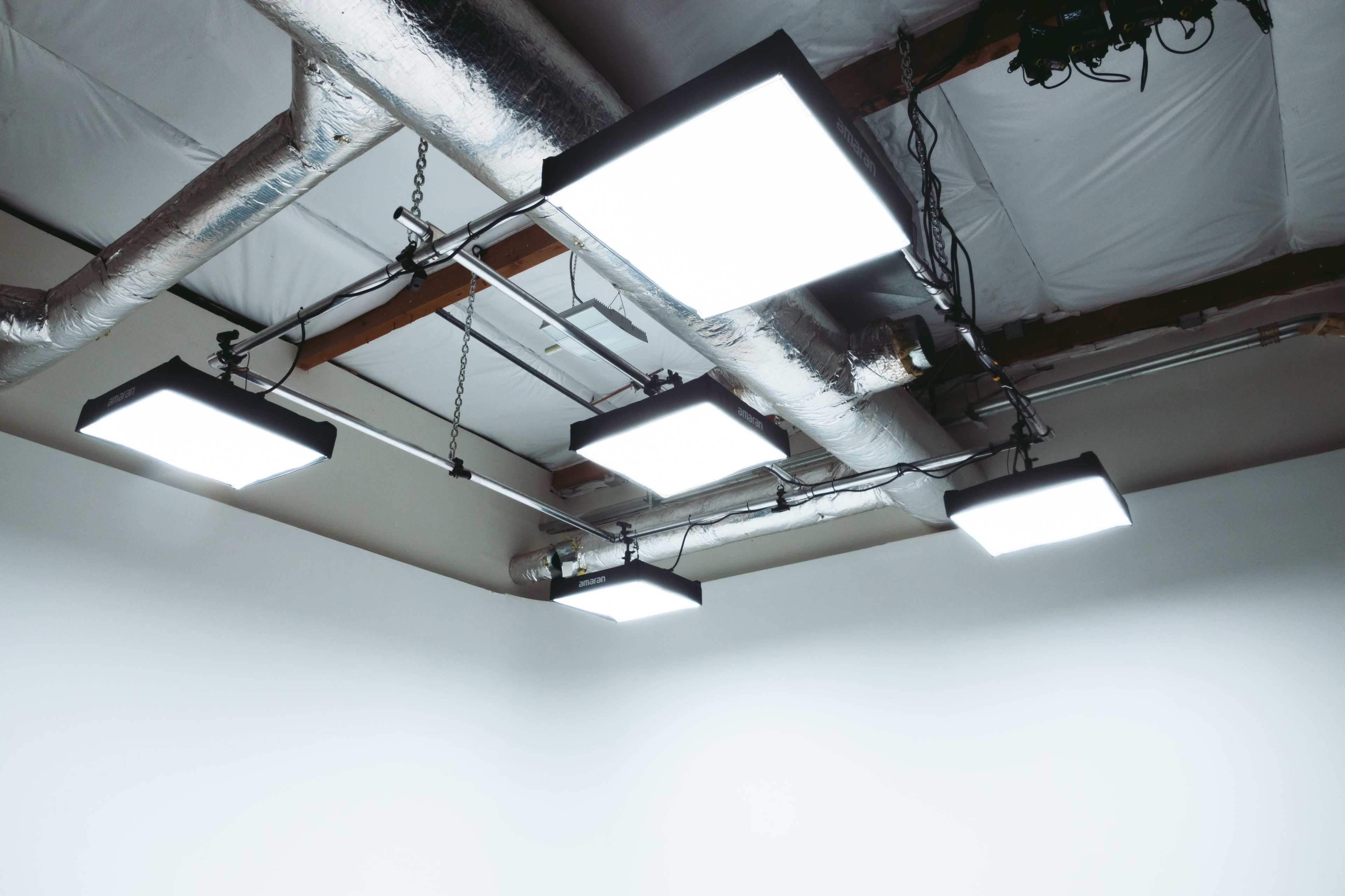 Five rectangular ceiling lights are mounted above a white wall in a room with exposed ductwork.