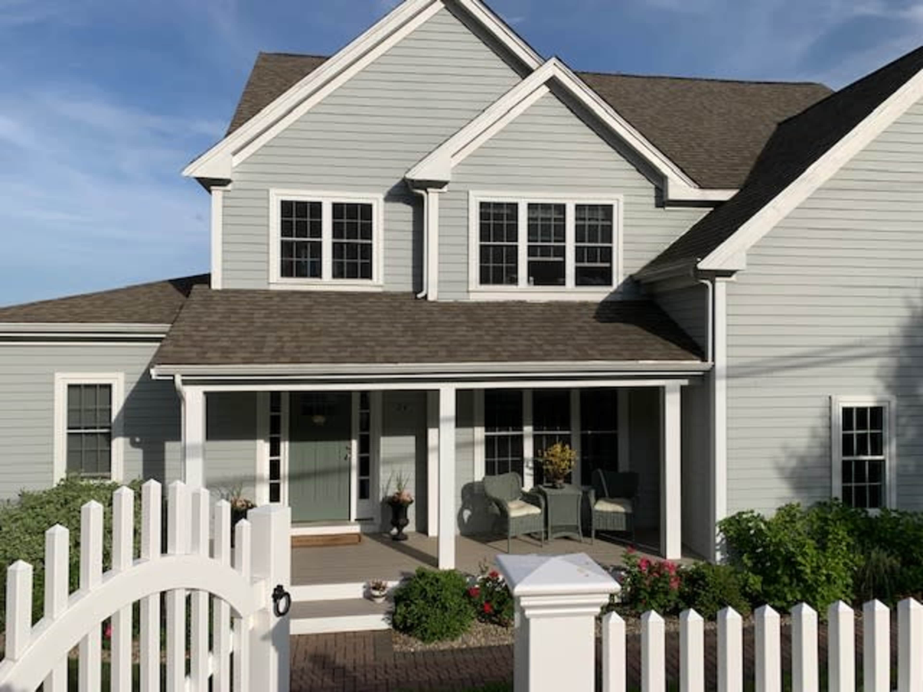 A light blue house with a covered porch and white picket fence features multiple windows and patio furniture.