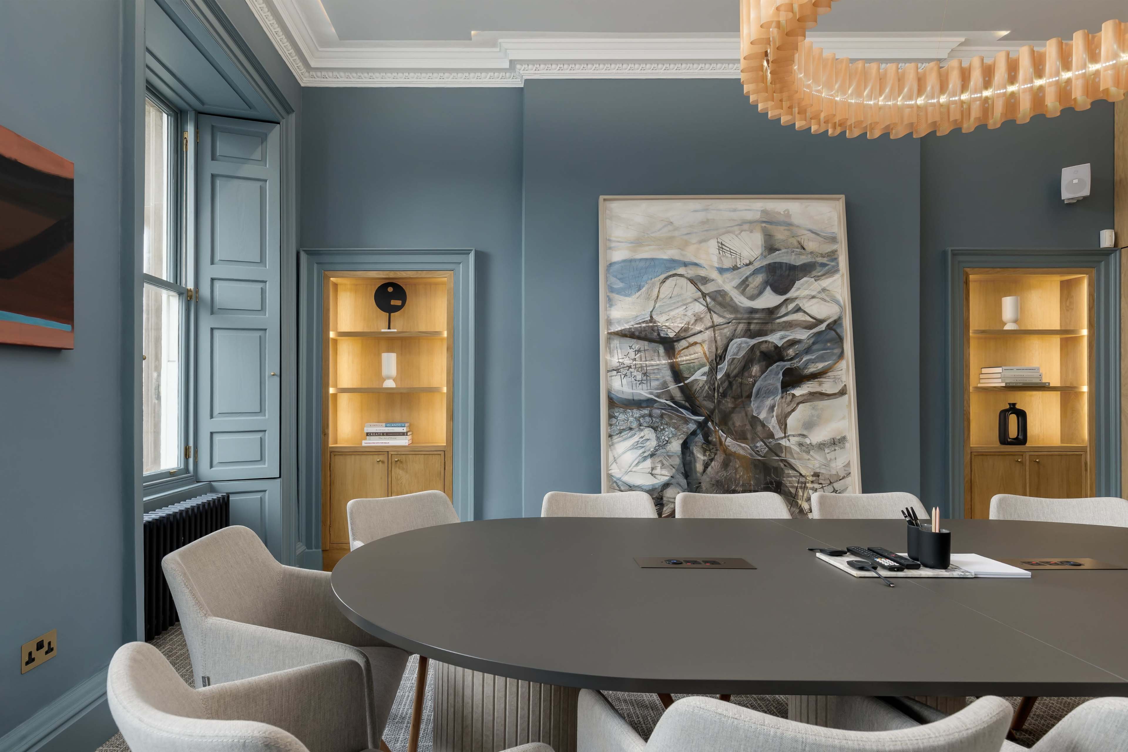 A modern conference room features a large oval table surrounded by upholstered chairs, with blue walls, a statement chandelier, and a large abstract painting.