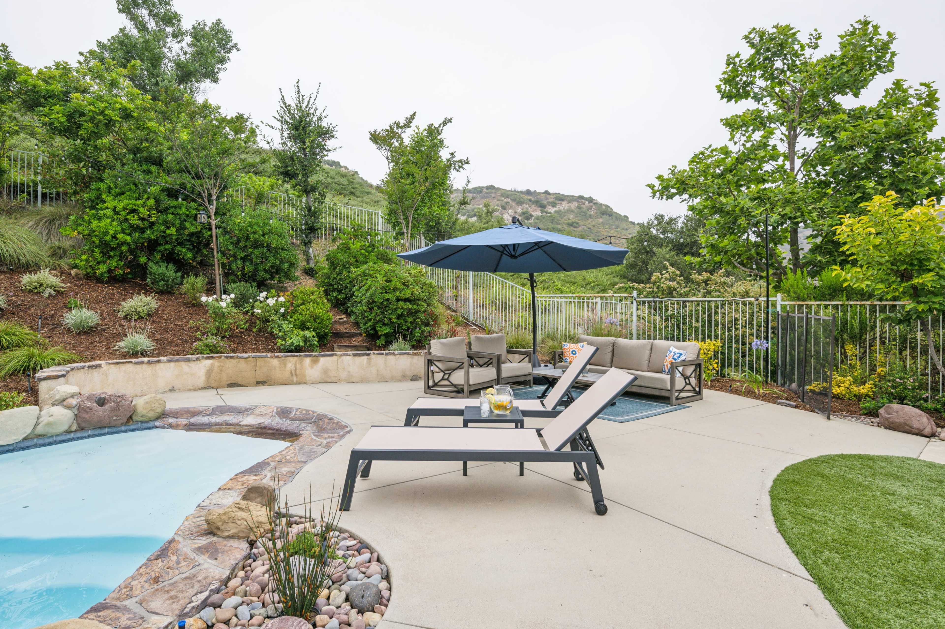 A patio area features two lounge chairs by a pool, surrounded by landscaped greenery and a shaded umbrella.