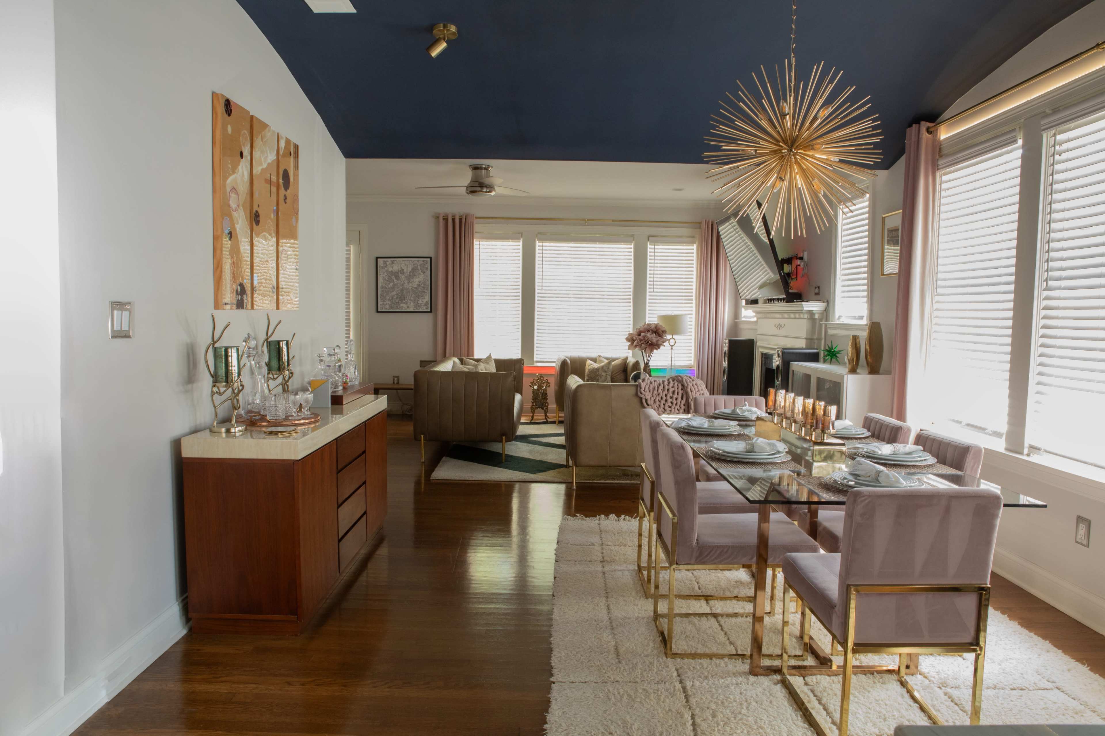 The image shows a modern dining area with a glass table set for a meal, surrounded by purple chairs, adjacent to a cozy living room featuring light-colored furniture and large windows.