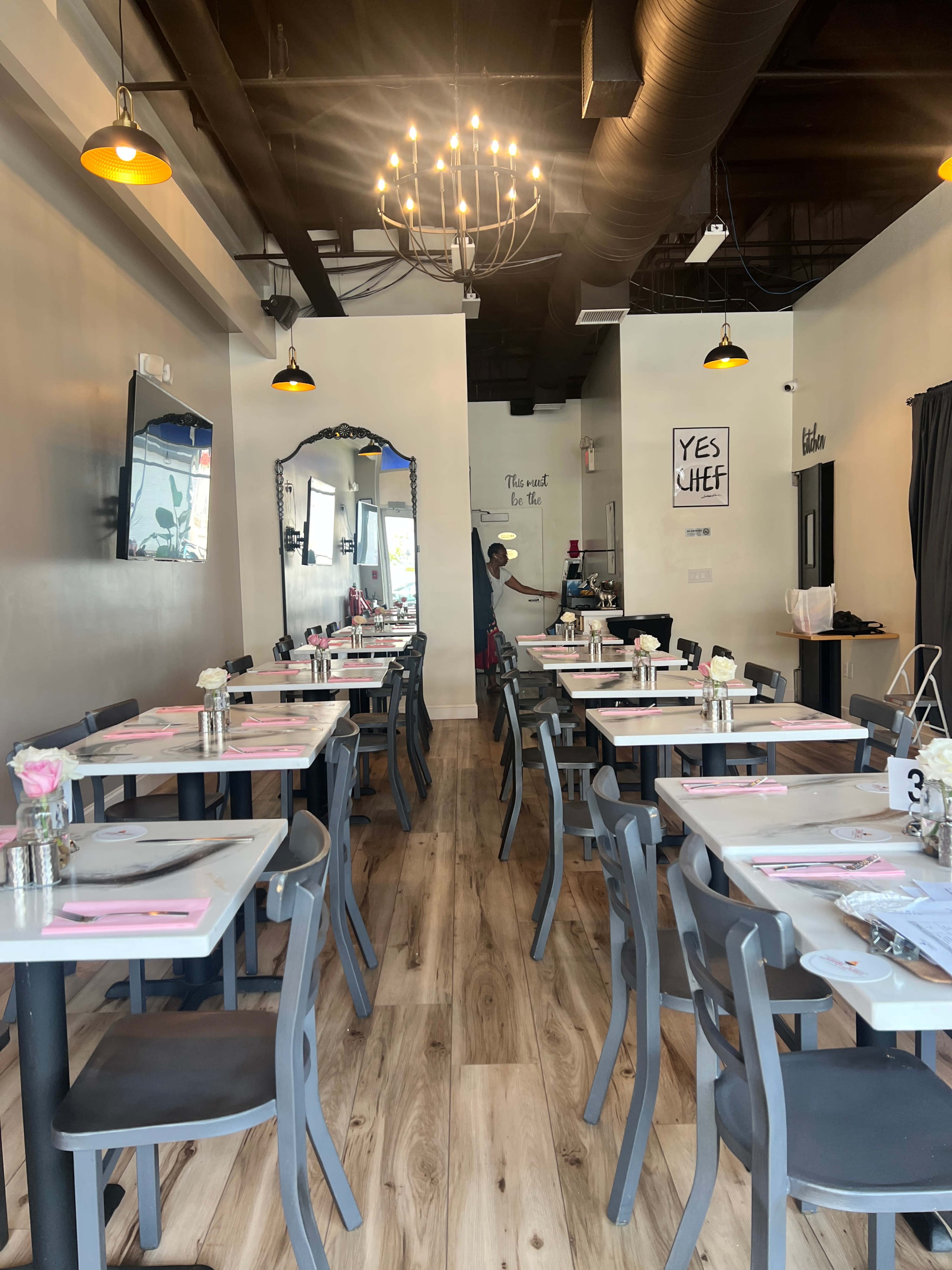 The image shows a modern restaurant interior featuring neatly arranged tables with pink placemats, floral centerpieces, and a decorative chandelier overhead.