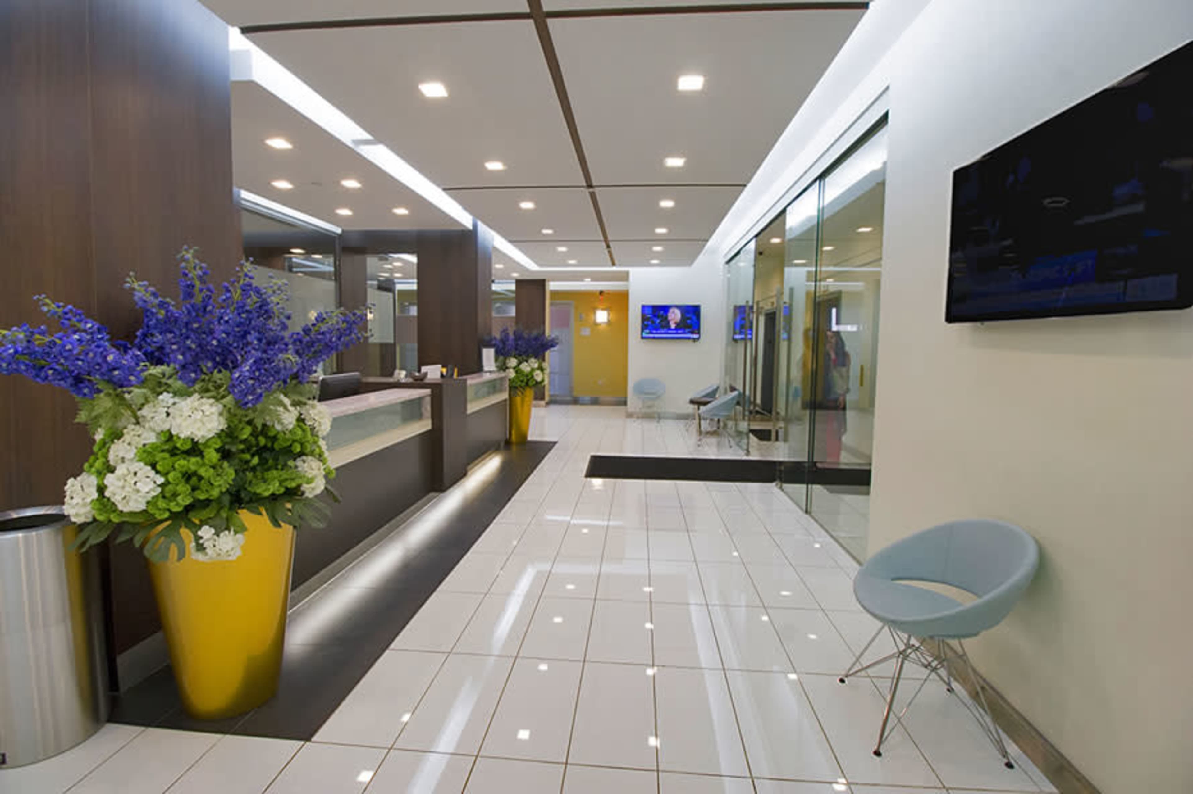 A modern reception area featuring a reception desk, a large floral arrangement in a yellow planter, and a television mounted on the wall.