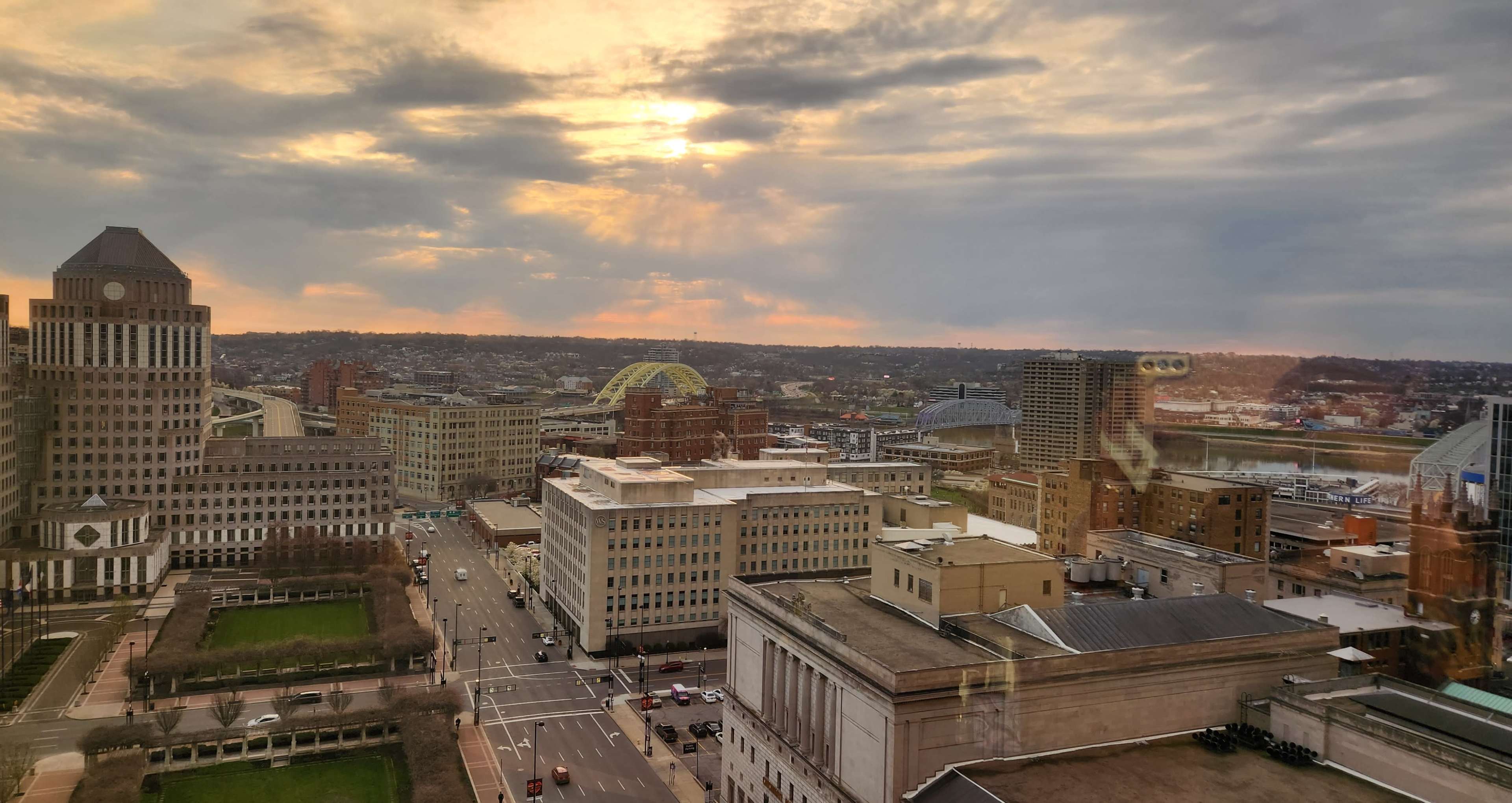 The image shows a cityscape at sunset, with buildings, streets, and a large bridge visible in the distance.