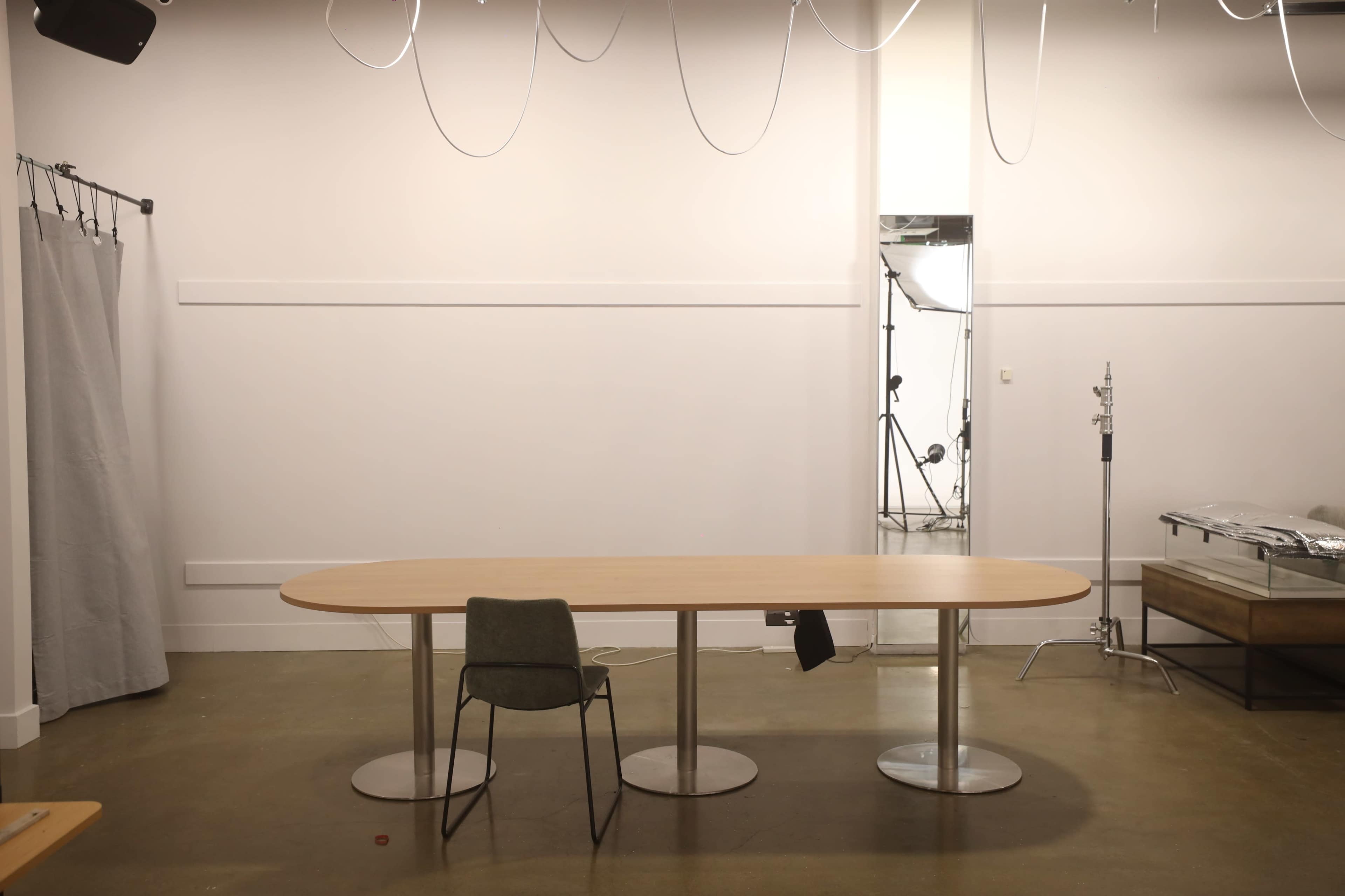 The image shows a minimalist room with a wooden oval table and three chairs, accompanied by studio lighting and equipment in the background.