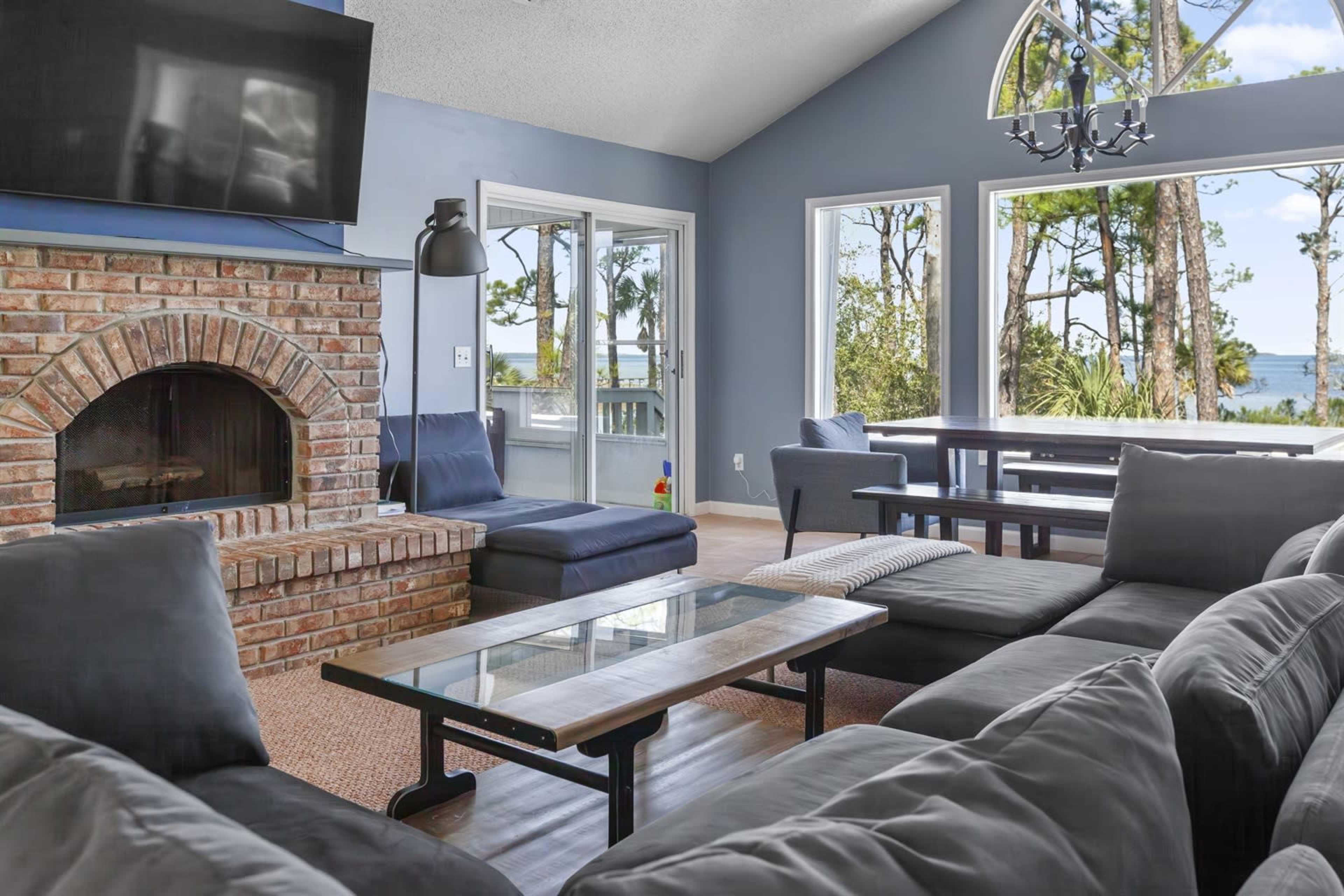 The image shows a spacious living room featuring a brick fireplace, large windows overlooking trees by a lake, and comfortable seating arranged around a coffee table.