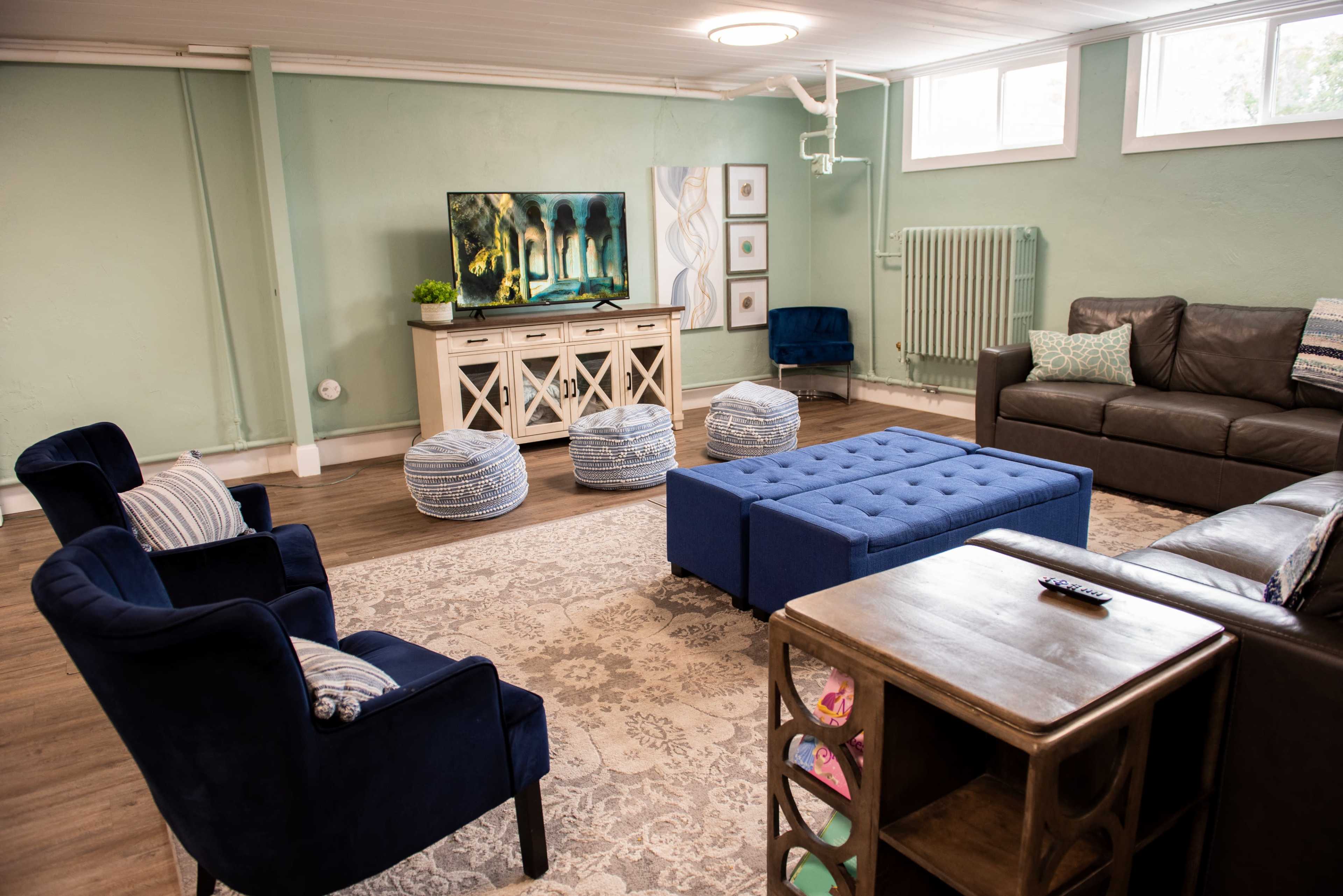 A cozy living room features a blue tufted ottoman, navy chairs, a brown sofa, a television on a white console, and patterned rugs.