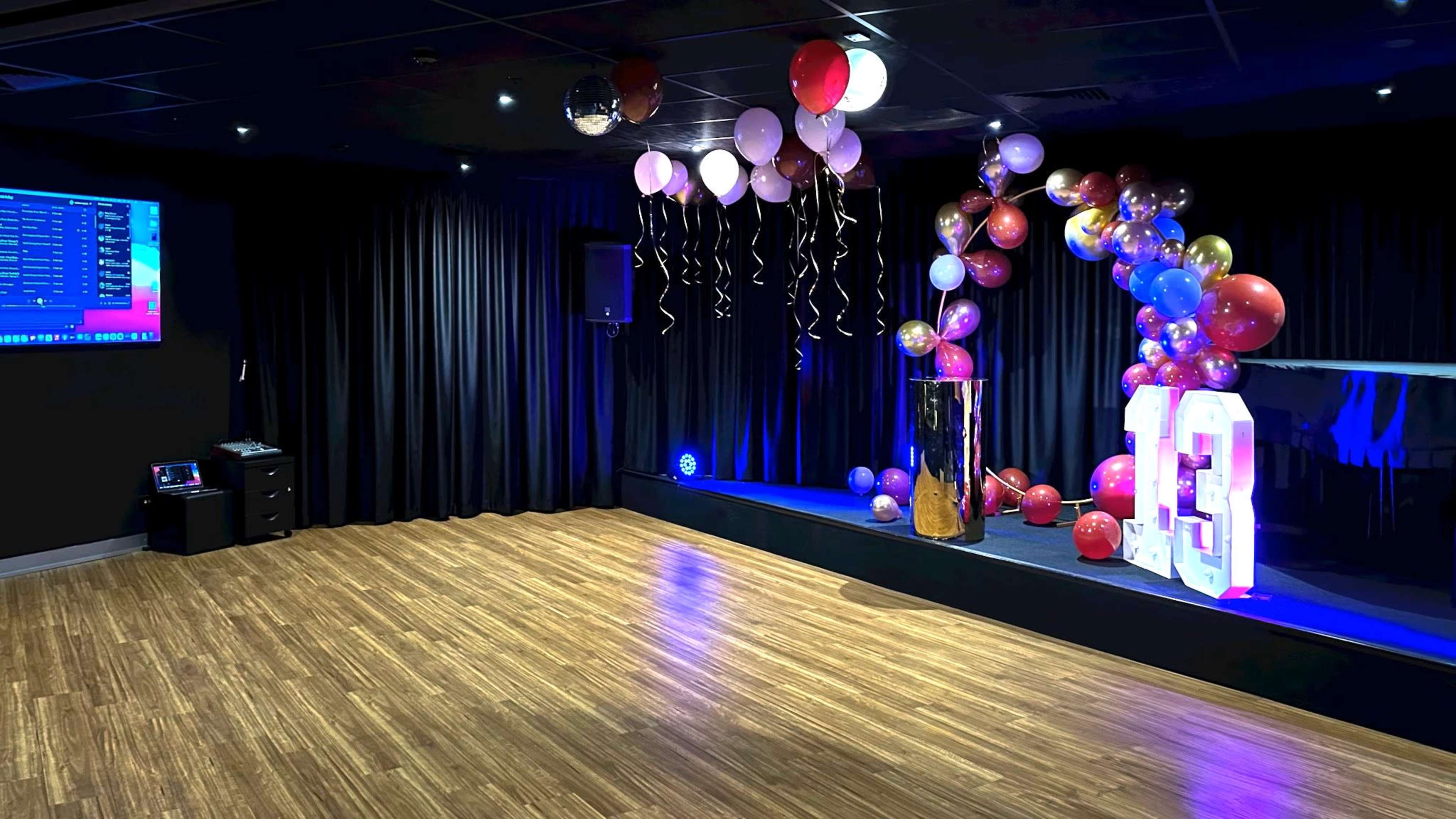 The image shows a sparsely decorated party venue with a stage adorned with colorful balloons and a large "13" display, set against a backdrop of black curtains and wooden flooring.