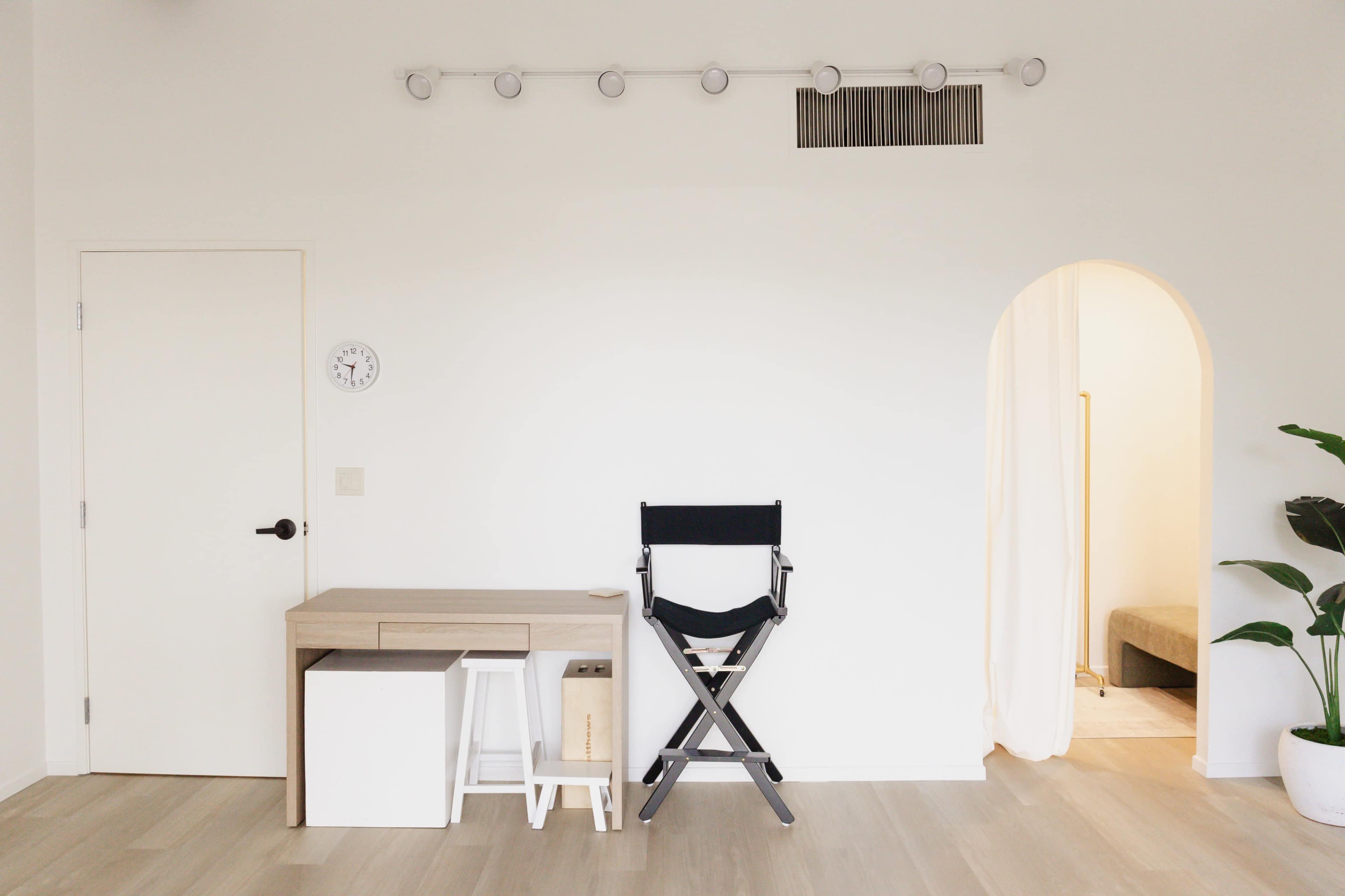 The image shows a minimalist interior with a desk, a director's chair, and a doorway leading to another room, all set against light-colored walls and wooden flooring.