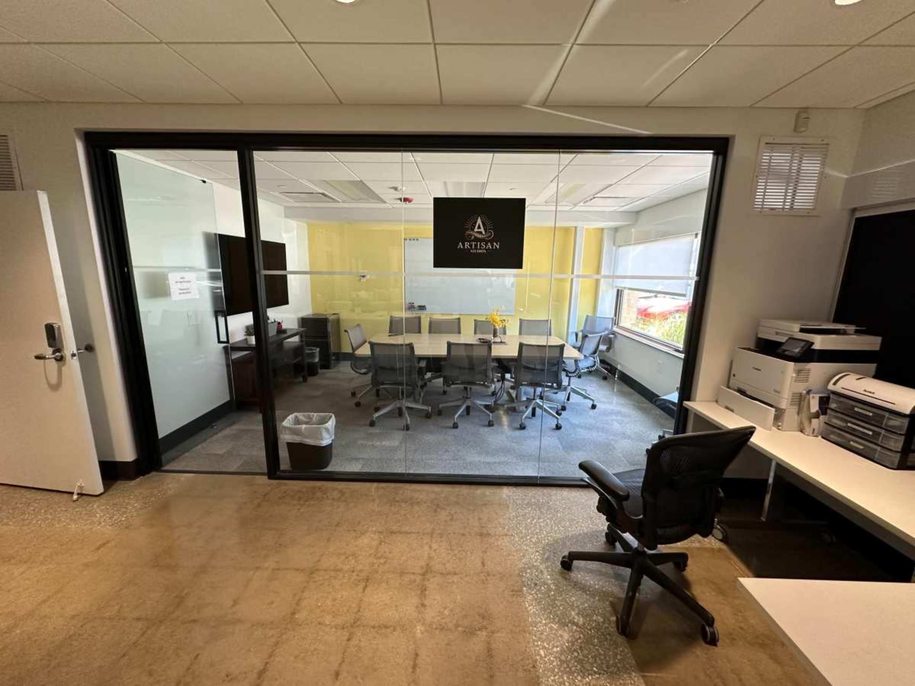 An office space with a glass-walled conference room featuring a large table and rolling chairs, accompanied by desks and office equipment outside the room.