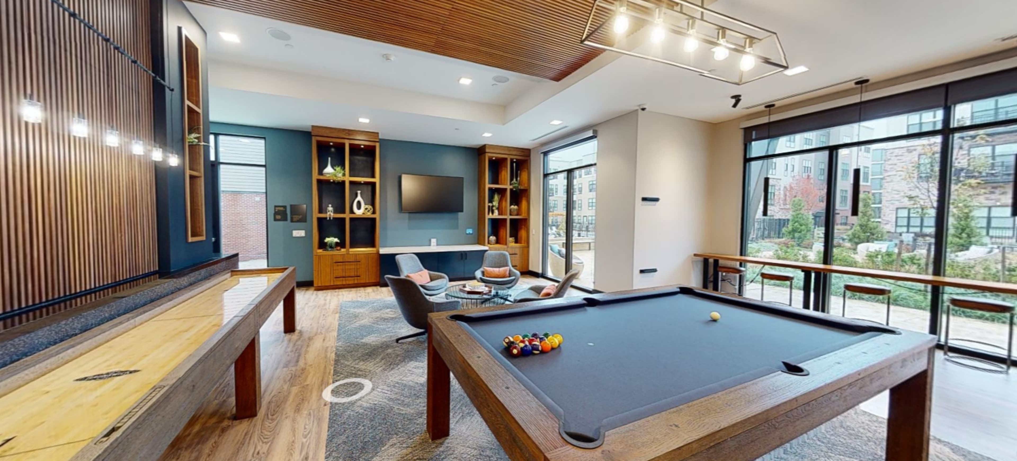 The image shows a modern recreational room with a billiard table, seating area, and large windows overlooking a garden.