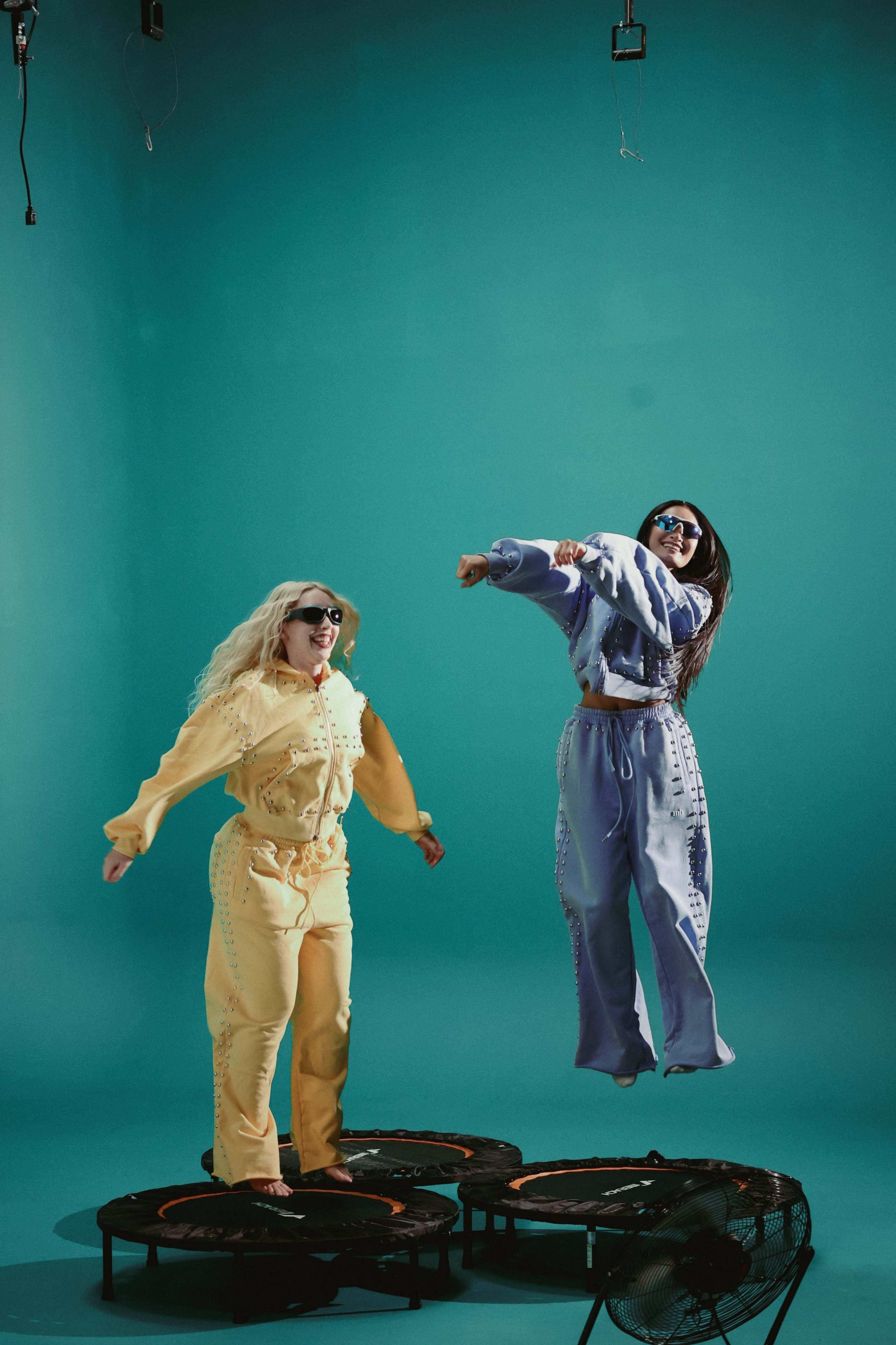 Two women in colorful tracksuits jump on mini trampolines against a teal backdrop.
