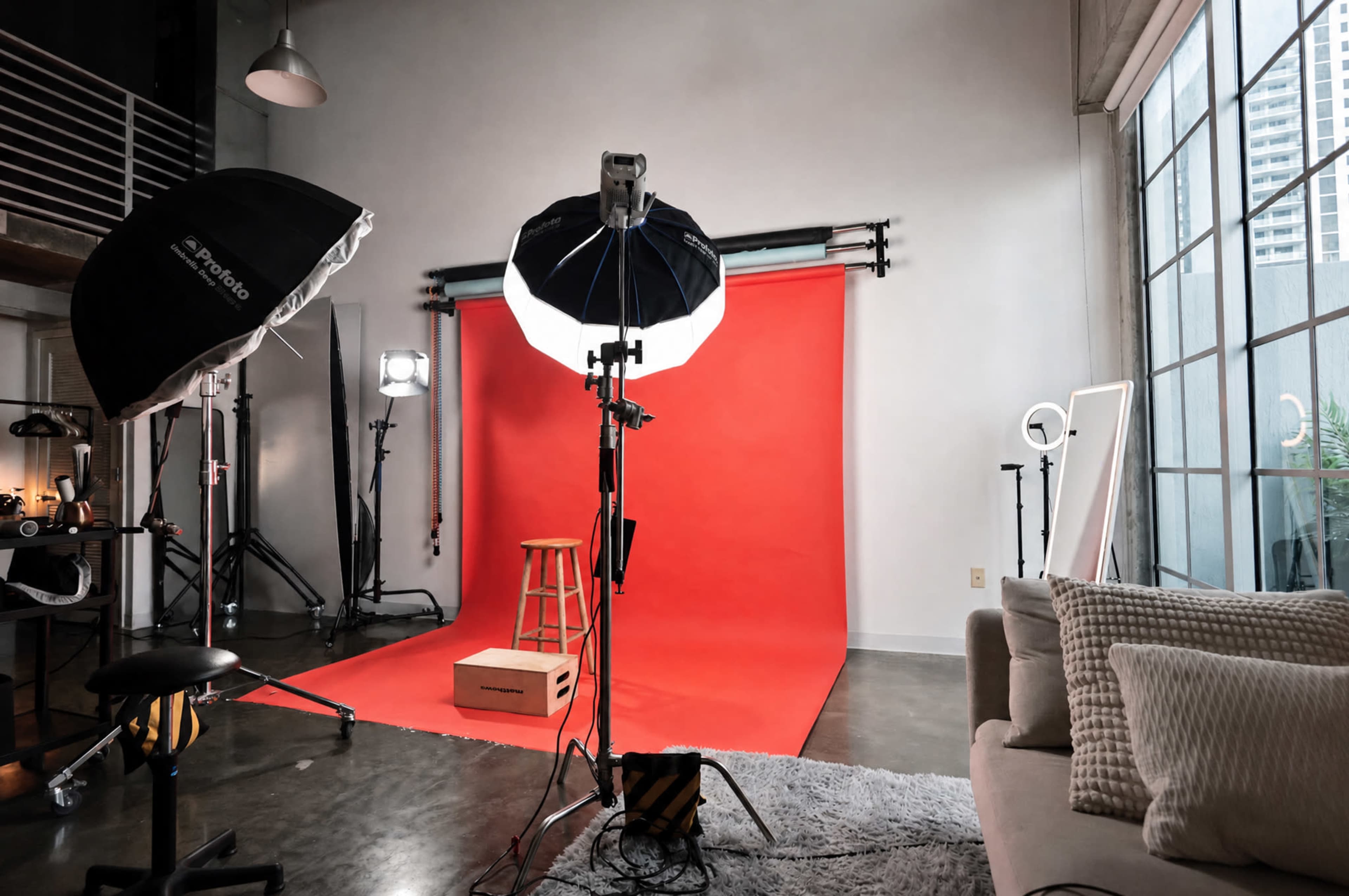 The image shows a photography studio set up with a red backdrop, a stool, and various lighting equipment.